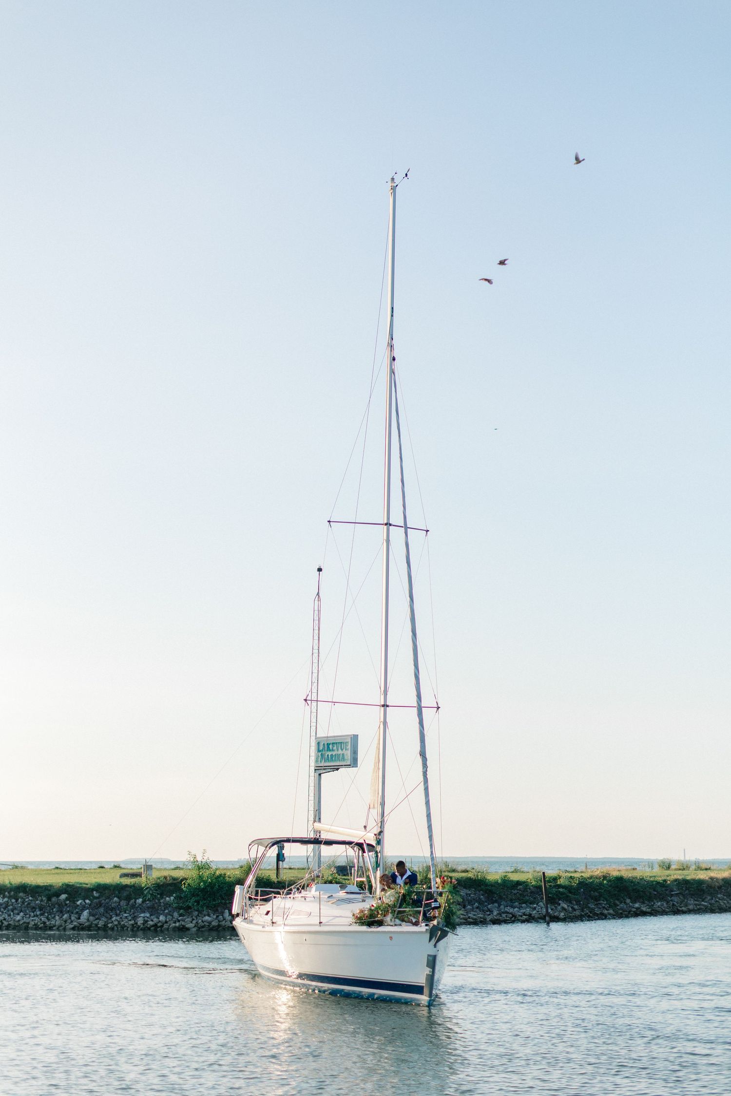 White sailboat gliding through calm coastal waters under a clear sky with birds flying overhead.