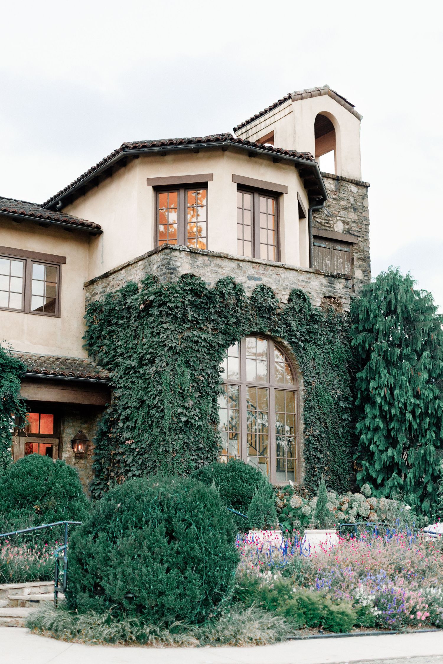 Elegant ivy-covered stone mansion with arched windows, manicured gardens, and a charming bell tower against a cloudy sky.