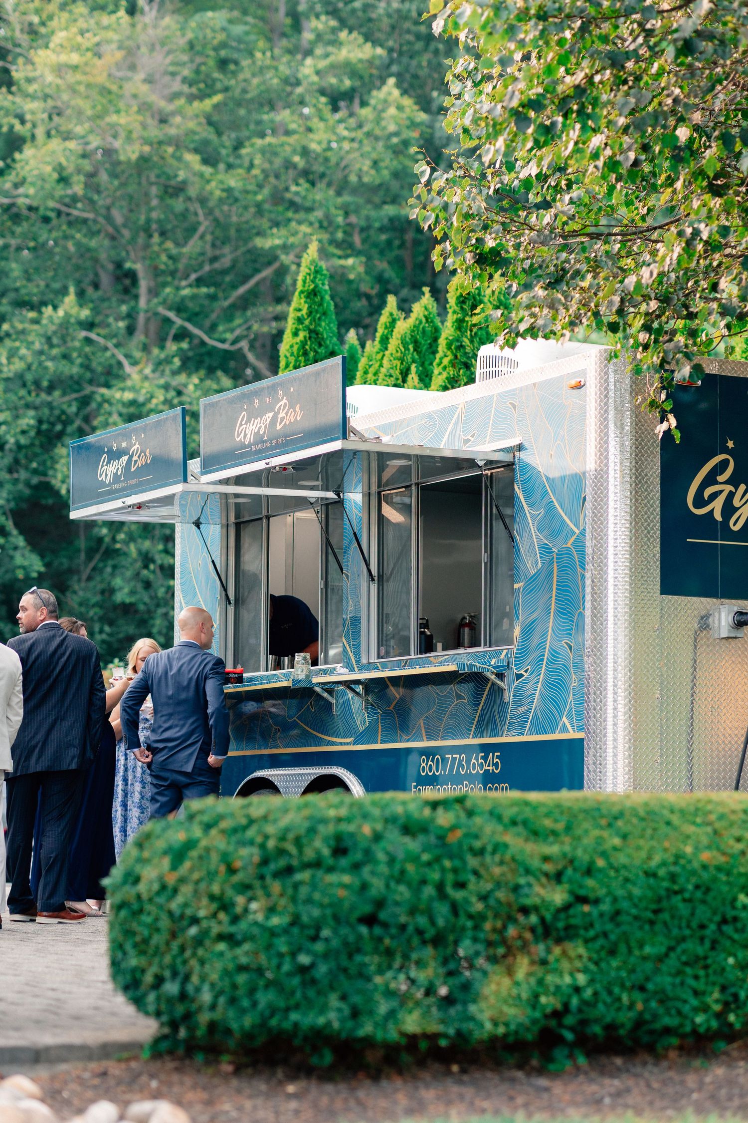 Elegant blue food truck trailer serving guests outdoors at an event, surrounded by lush green trees and manicured hedges.