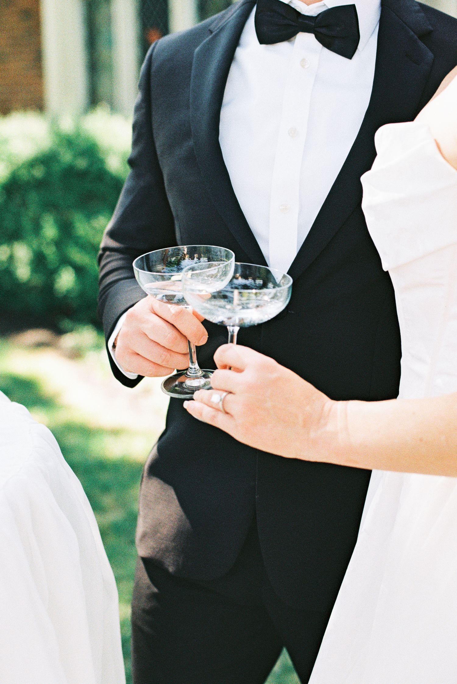 Bride and groom in classic black tuxedo and white gown toasting with vintage coupe champagne glasses outdoors.