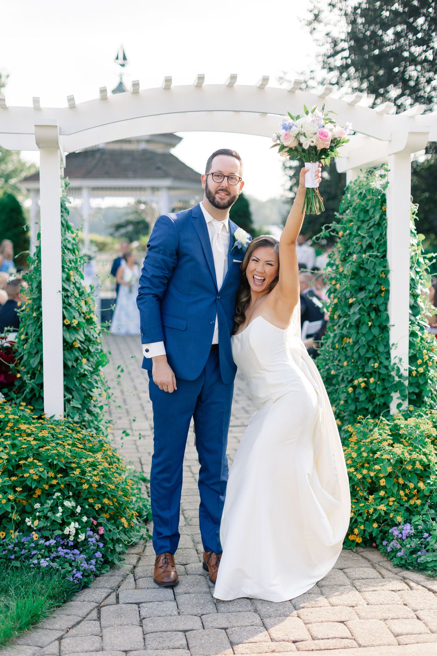 Joyful bride in white gown raises bouquet beside groom in blue suit under white pergola in lush garden.