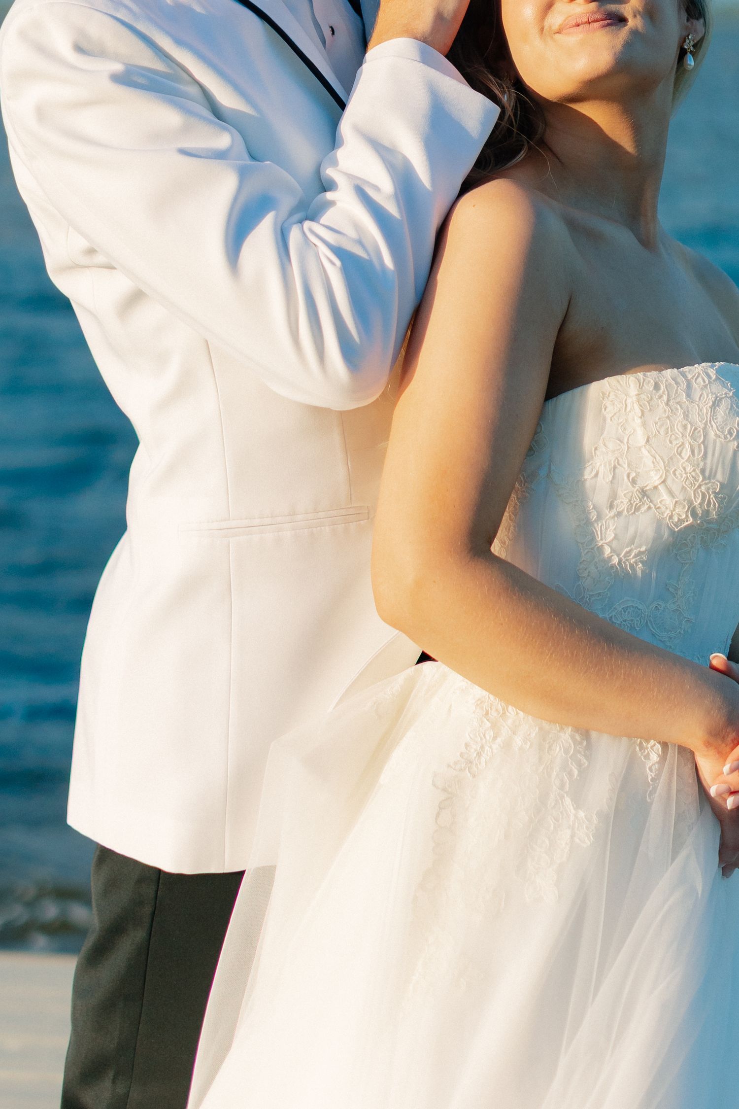 Bride and groom in white attire embrace on a beach with blue ocean water in the background, wedding at Bella Cosa