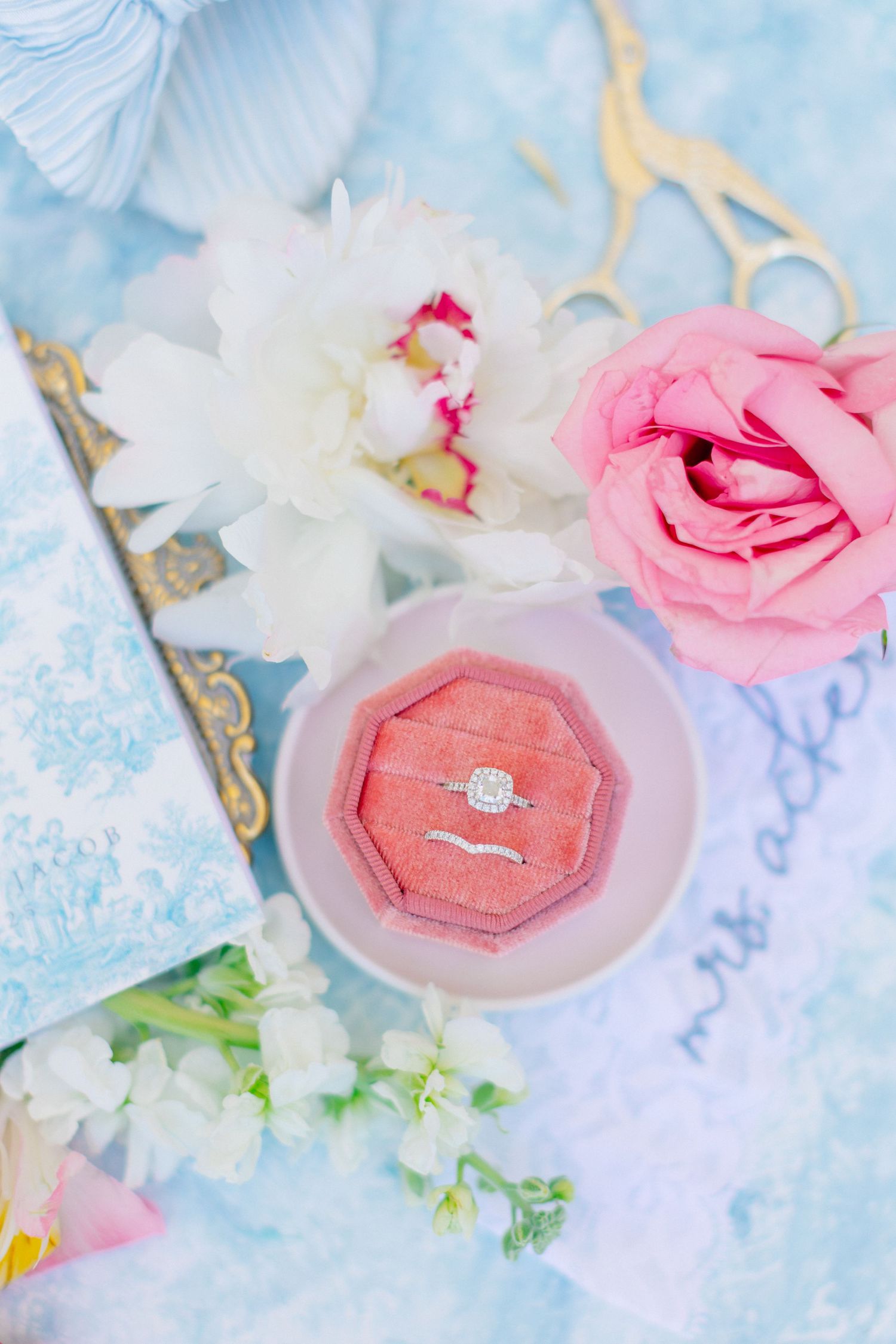Diamond engagement ring in pink velvet box surrounded by peonies and roses on blue background, at Bella Cosa in Florida