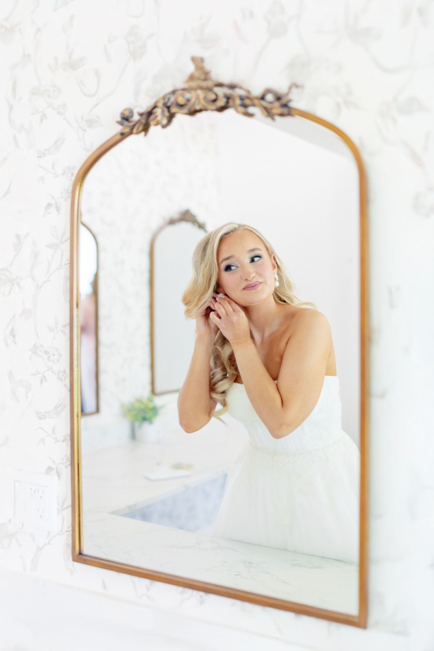 Blonde bride in white dress adjusting earring, reflected in ornate gold arched mirror with floral wallpaper background, wedding at Bella Cos