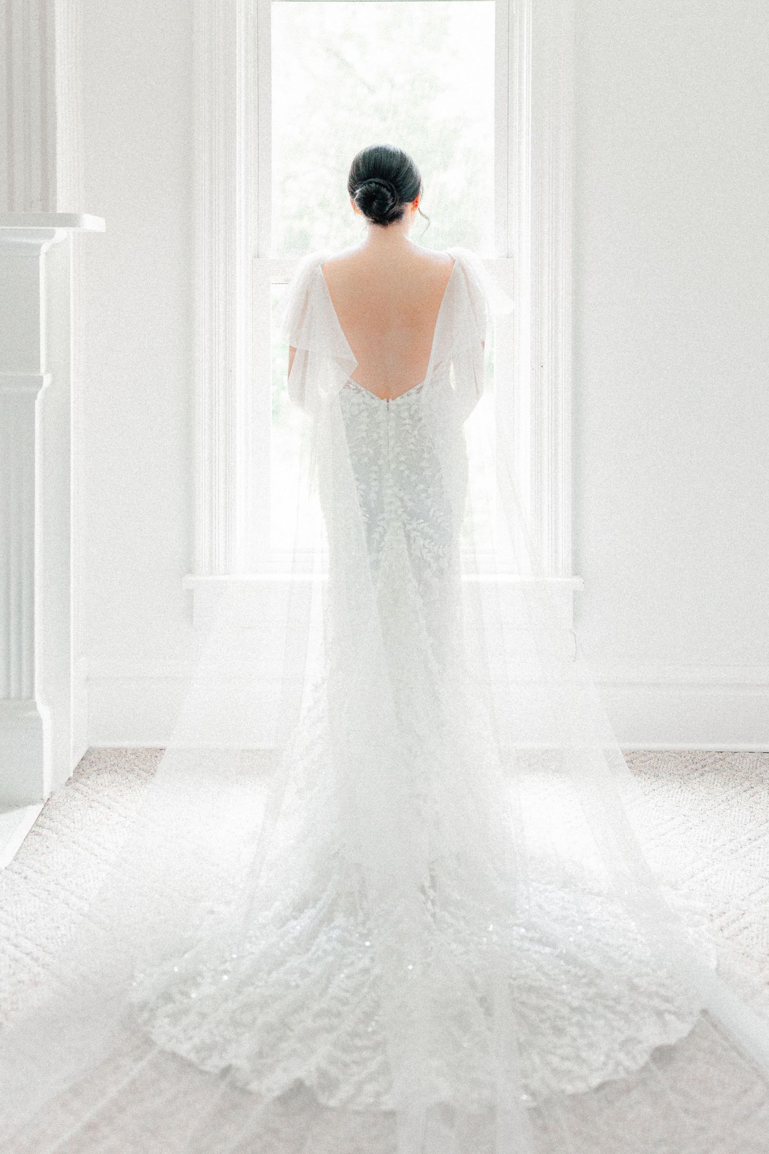 Bride in a stunning backless lace wedding gown with a long train stands before a bright window.