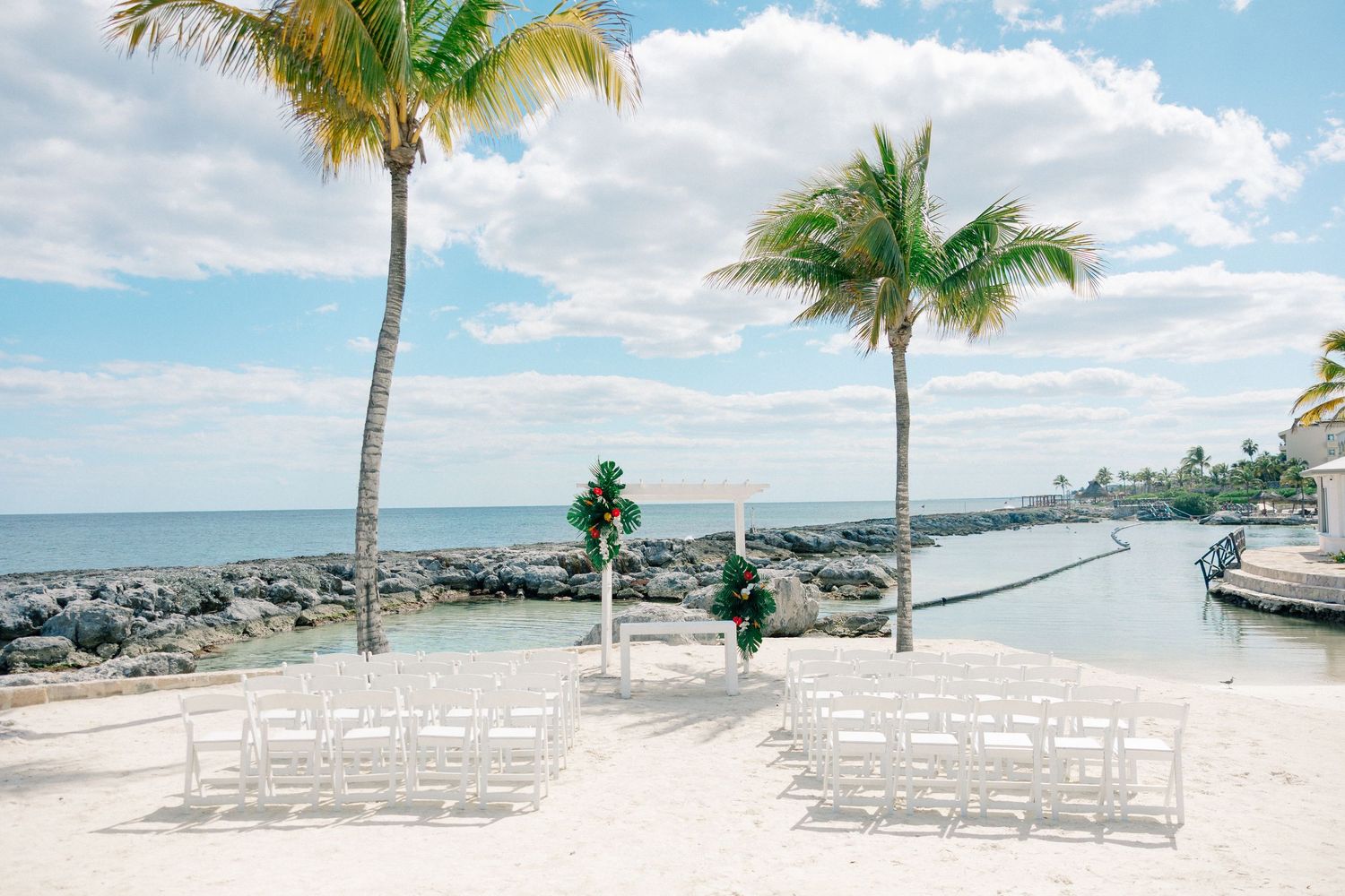 Outdoor beach wedding ceremony setup with white chairs, floral arch, palm trees, and ocean views under a bright blue sky.