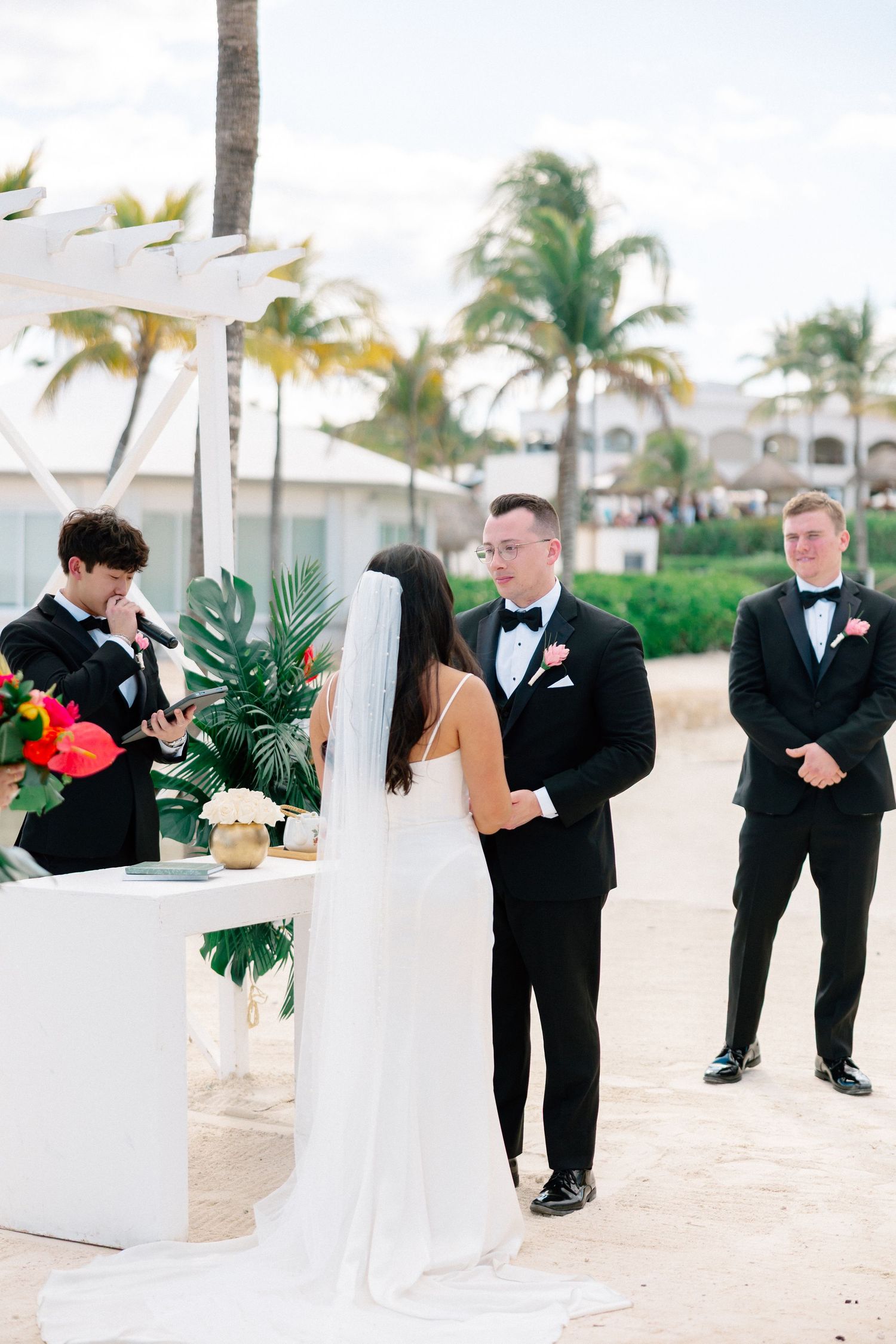 Bride and groom exchange vows at a tropical beach wedding ceremony with palm trees and white pergola.