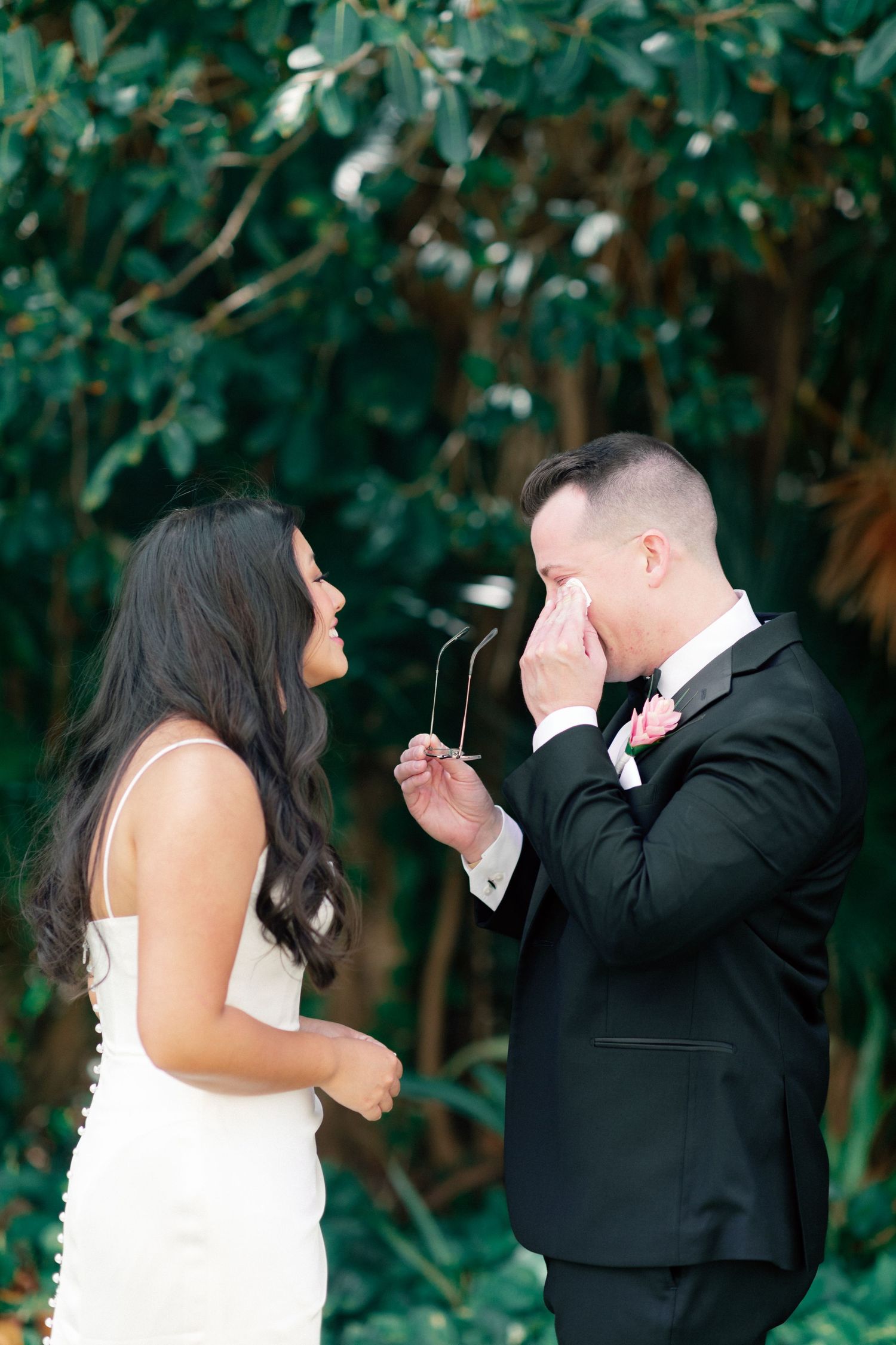 Emotional groom wiping tears while seeing bride for first time on wedding day, surrounded by lush greenery.