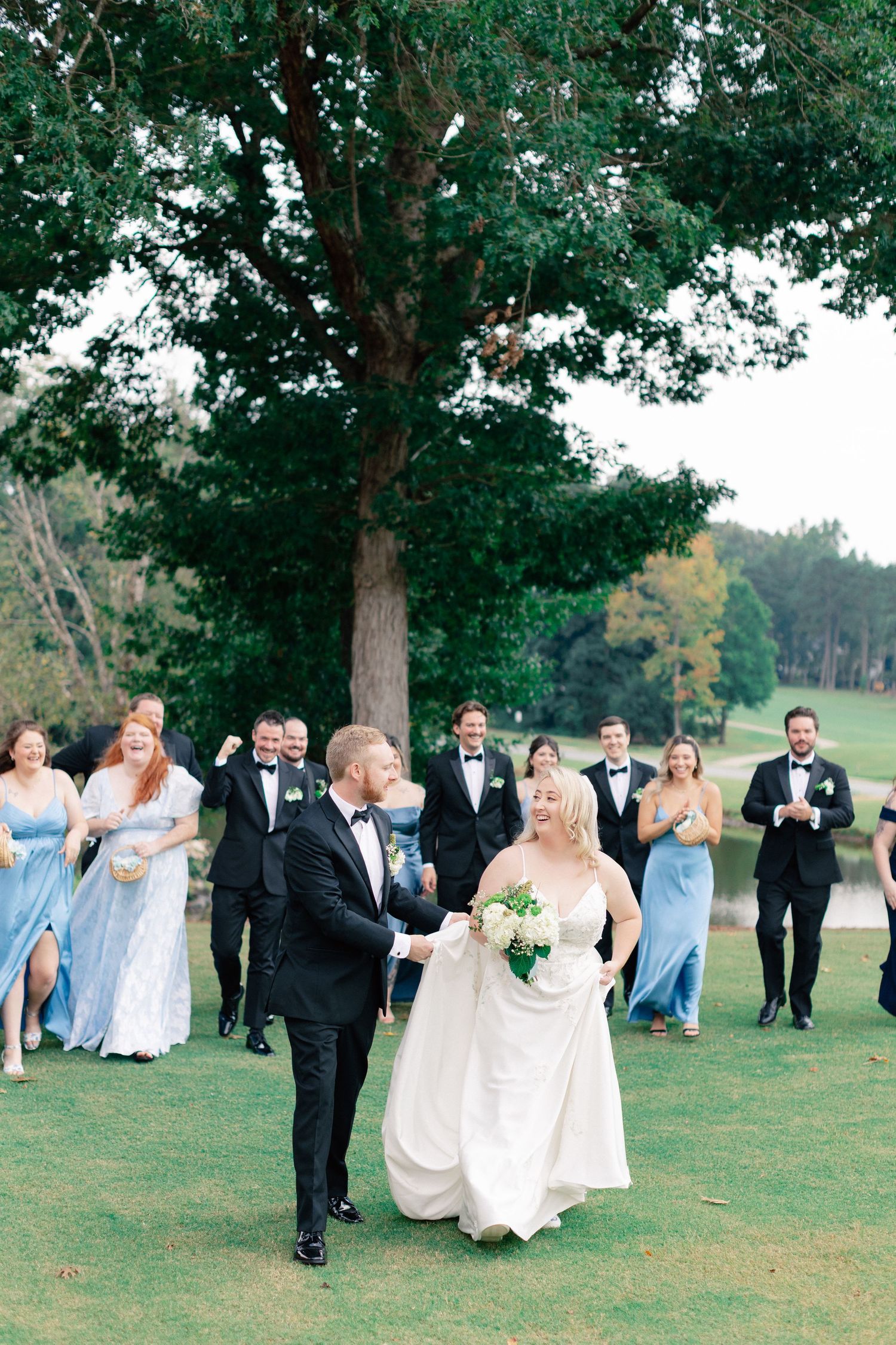 Bride and groom walk hand-in-hand on green lawn as wedding party in blue gowns and black tuxedos celebrate behind them.
