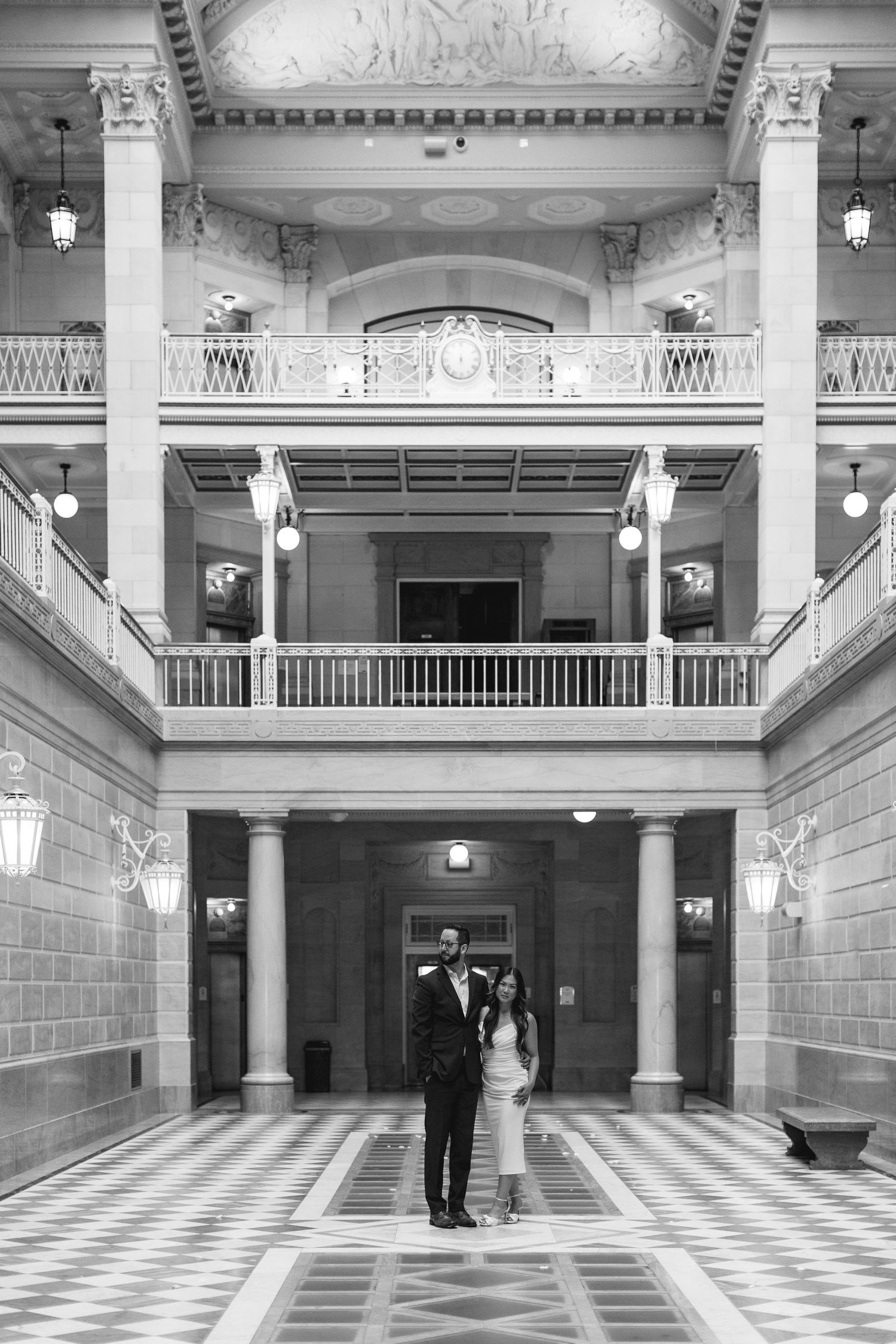 Black and white photo of a couple posing in an elegant grand hall with ornate balconies and checkered floors.