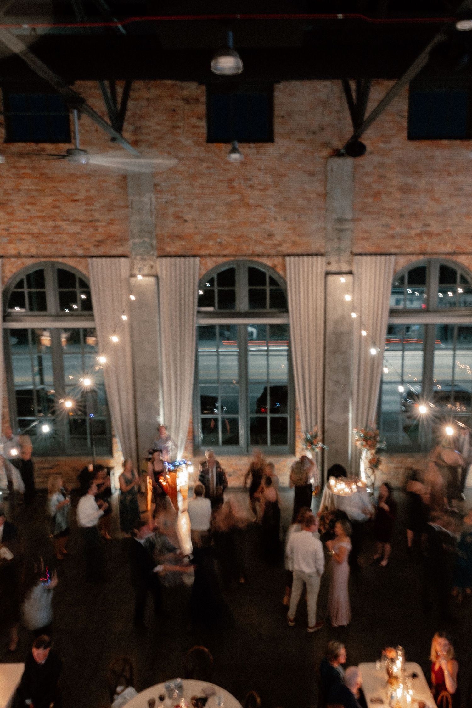 Aerial view of elegant wedding reception in industrial brick venue with arched windows, string lights, and guests dancing on the floor.