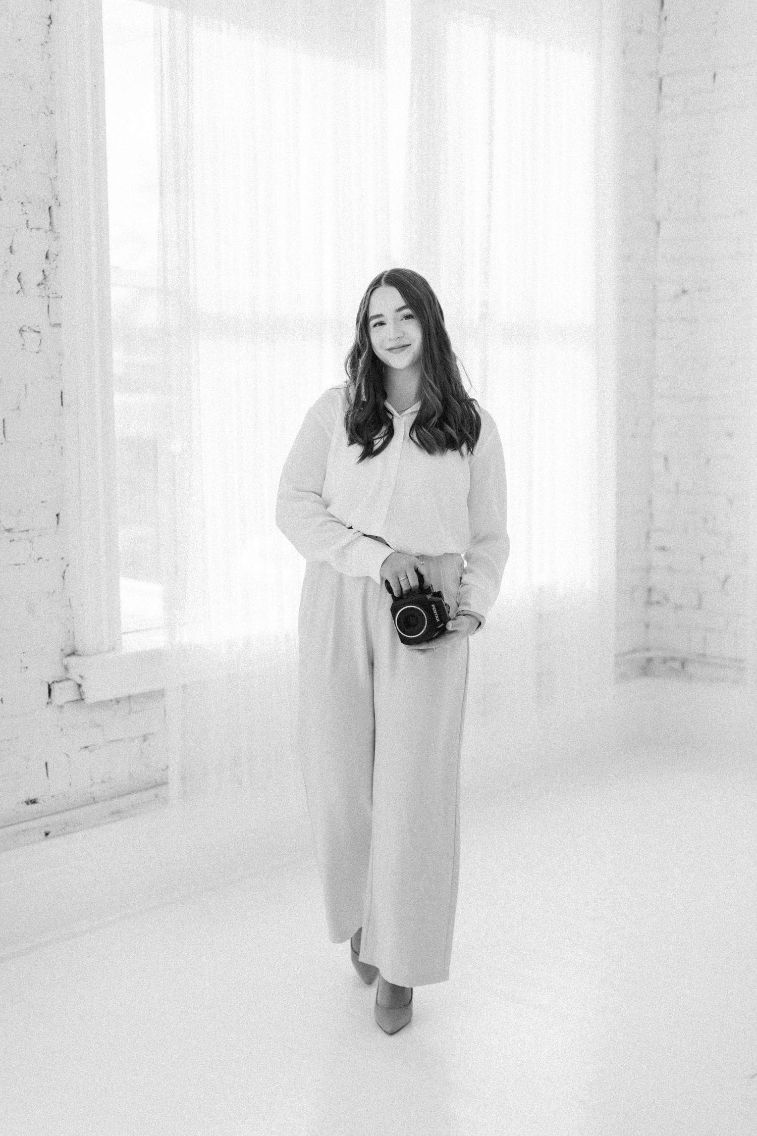 A woman in a white outfit holds a camera while standing in a bright white brick-walled studio, black and white photo.