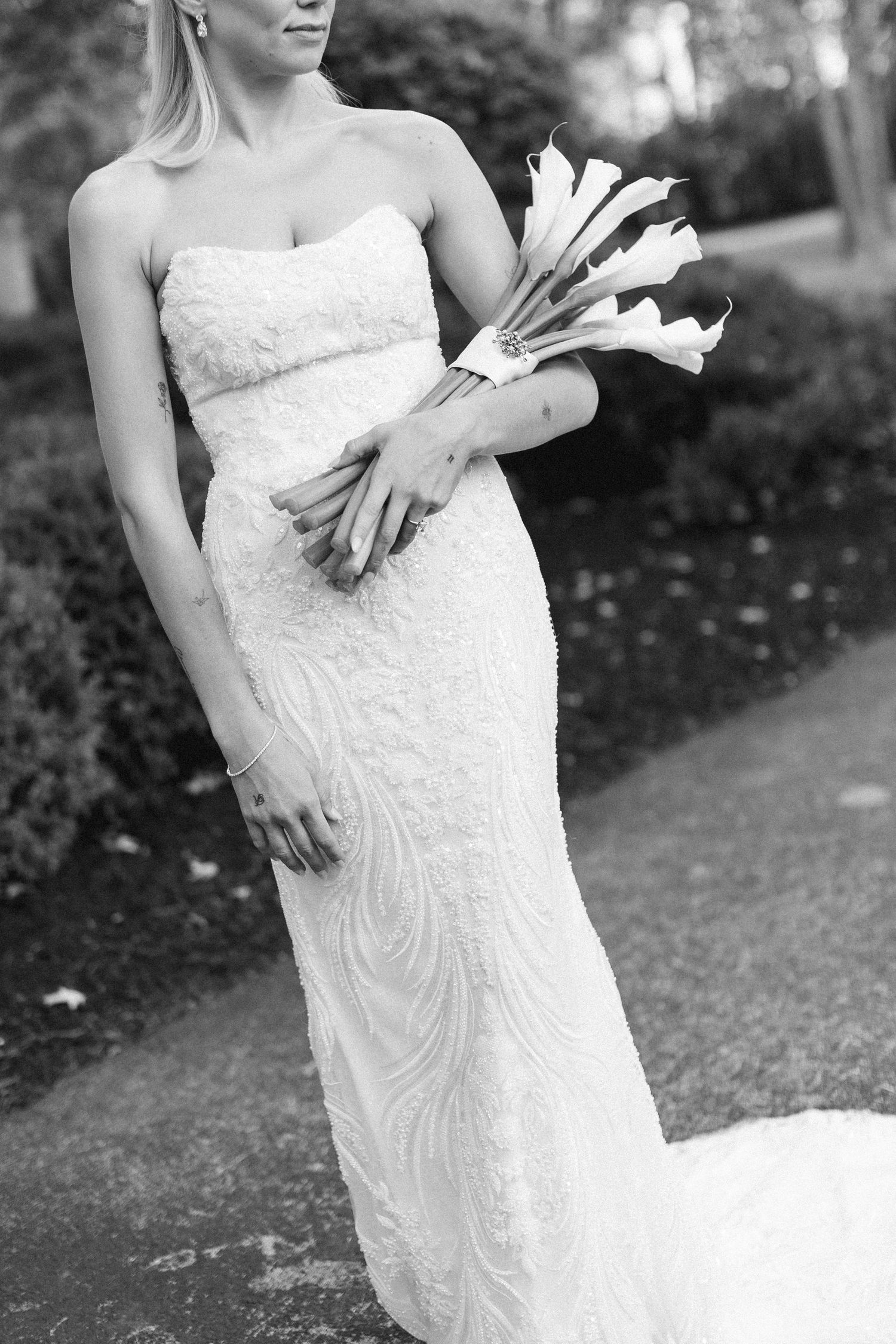 Black and white portrait of bride in fitted strapless lace gown holding elegant calla lily bouquet.