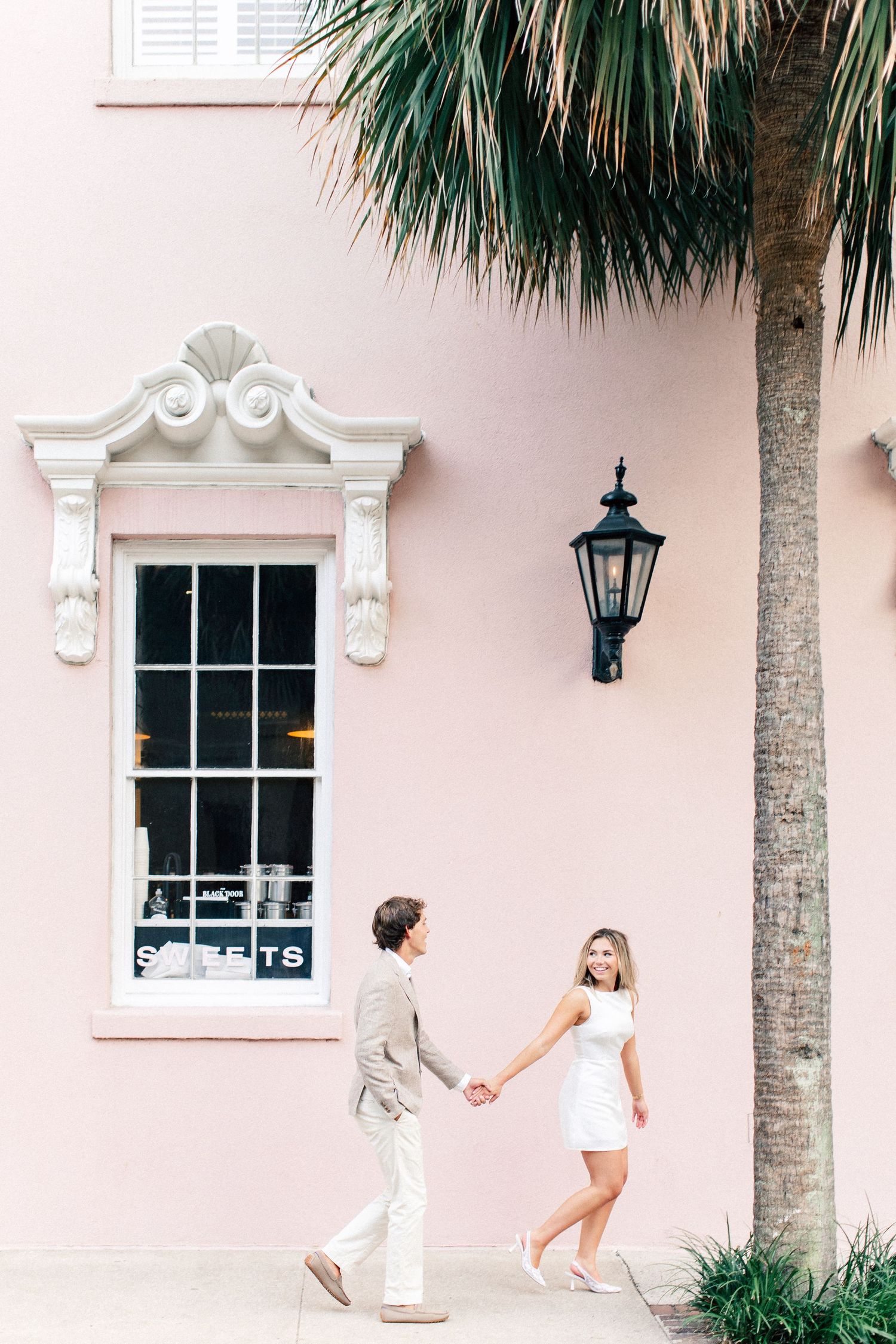 Couple holding hands walking along a pink historic building with ornate window trim and a palm tree in Charleston.