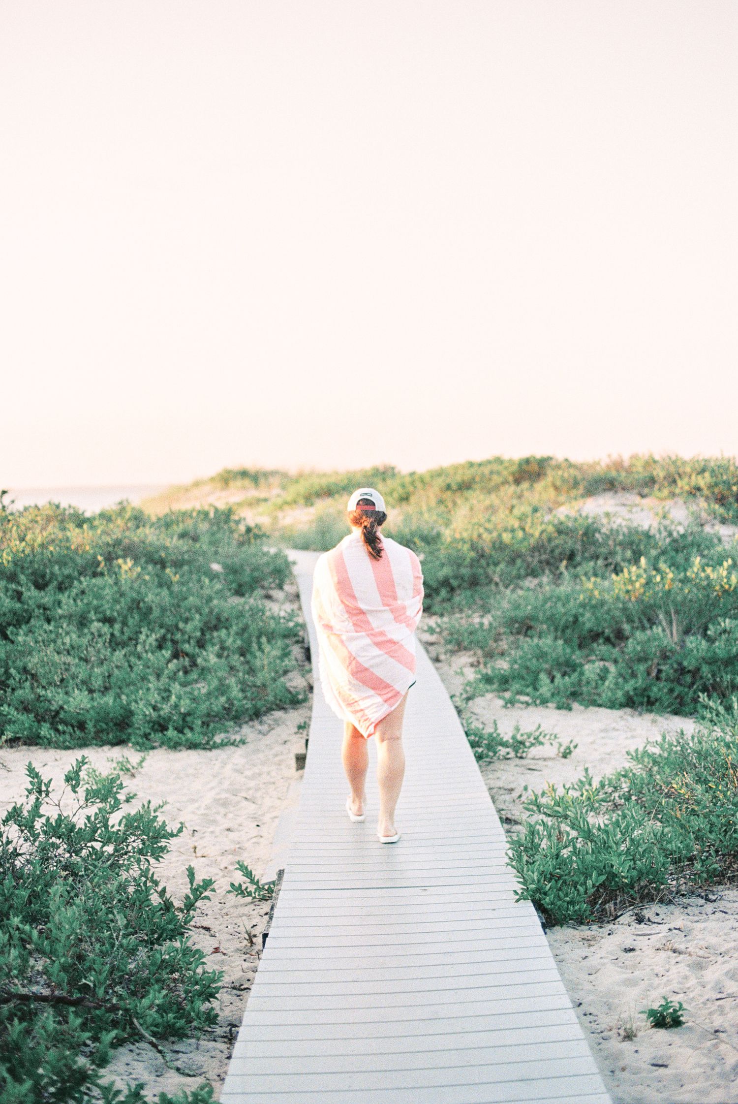 Woman wrapped in pink striped towel walking on wooden boardwalk path through coastal dunes at sunset.