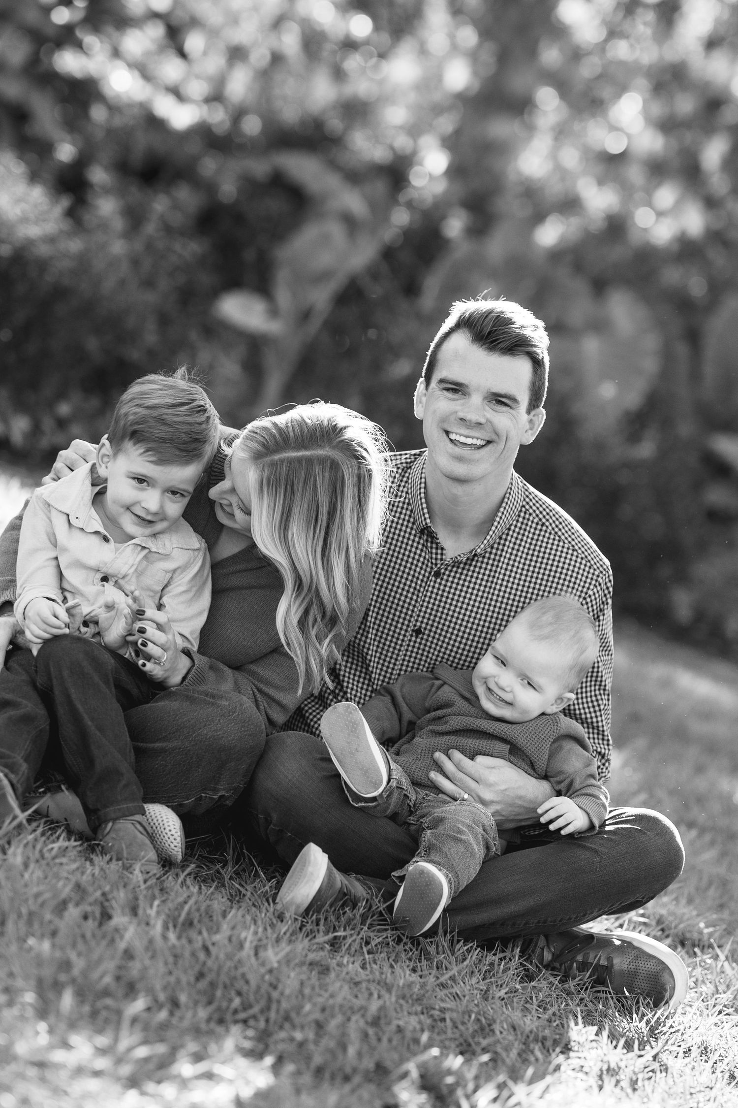 Black and white family portrait of parents with two young children sitting on grass outdoors.