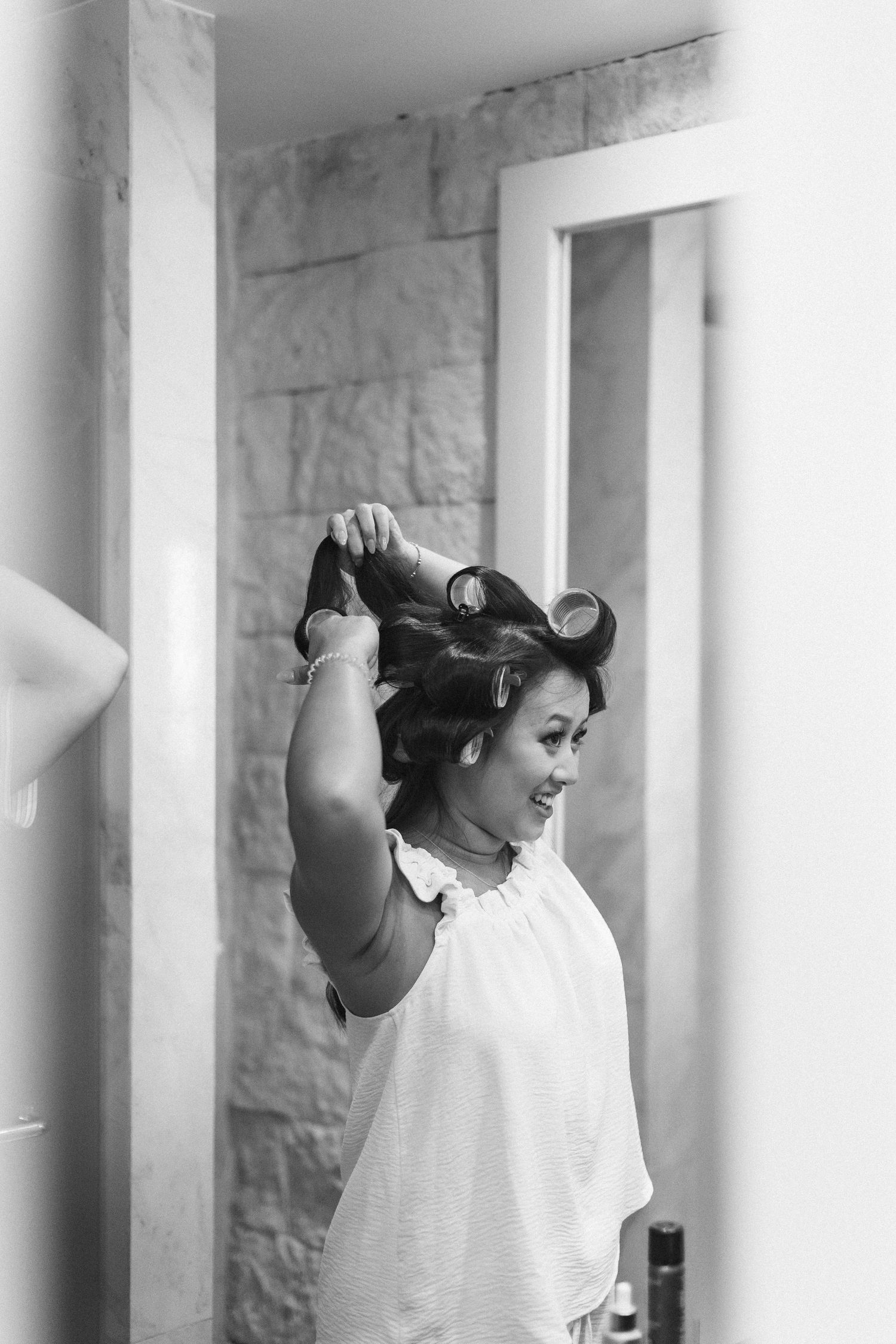 Black and white photo of a woman styling her hair with rollers, smiling in a mirror reflection.