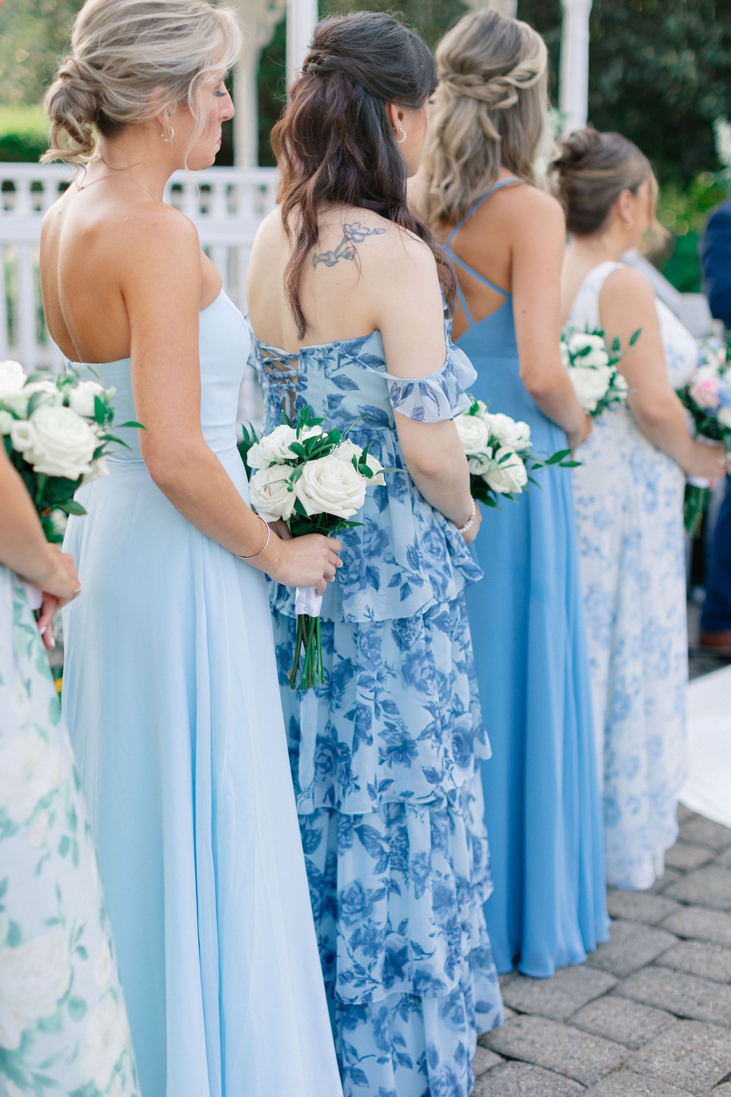 Bridesmaids in blue dresses holding white rose bouquets stand in a row at an outdoor wedding ceremony.