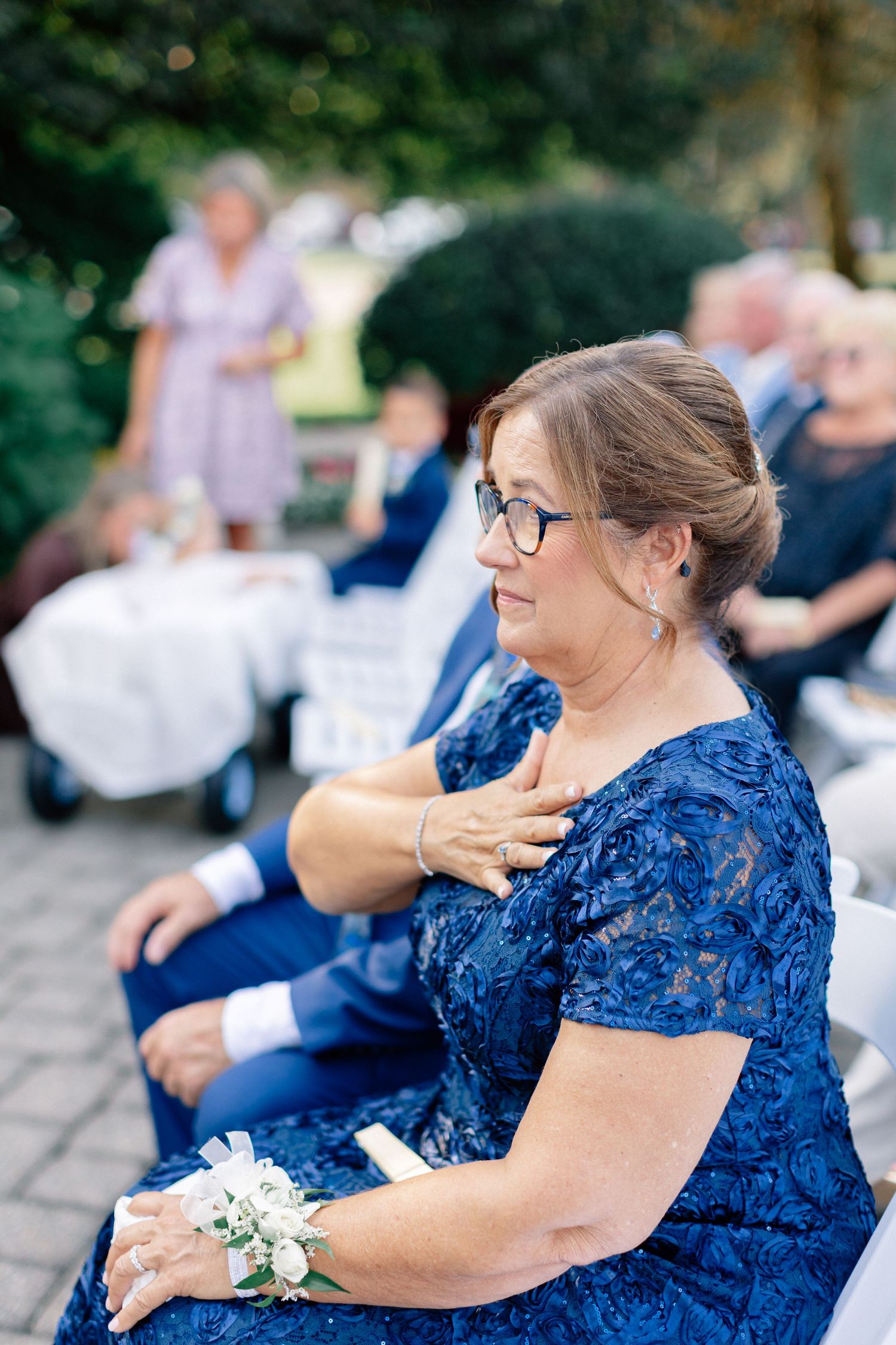 Woman in blue lace dress with wrist corsage seated at outdoor wedding ceremony, hand on chest.