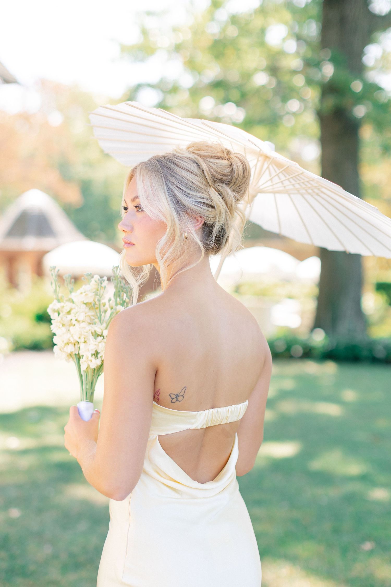 Bride with updo hairstyle and open-back strapless gown holding white parasol and flowers, showing back tattoo.