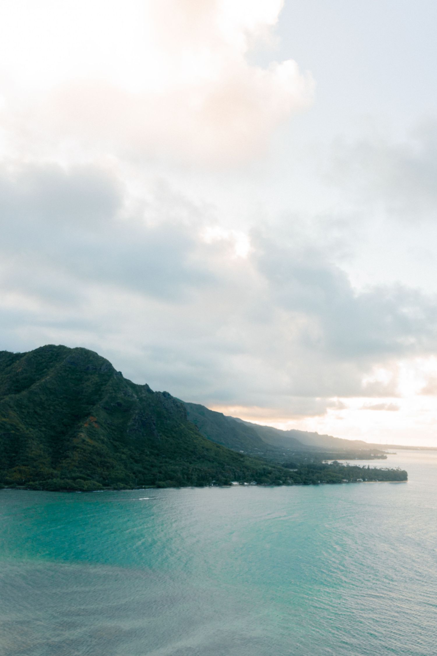 Aerial view of lush green mountain coastline with turquoise ocean waters under a cloudy sky in Hawaii.