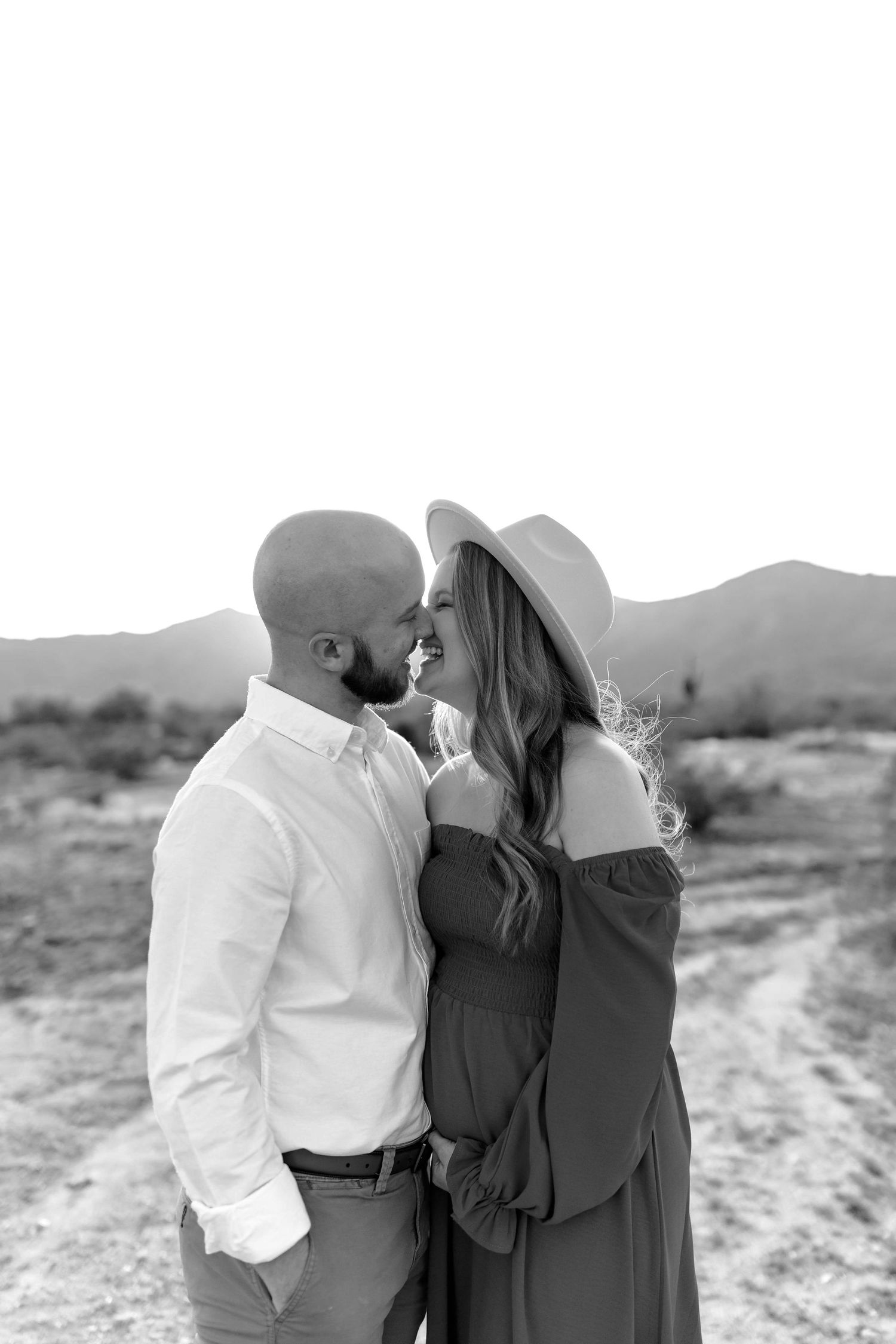 Black and white photo of a couple sharing a romantic moment in a desert landscape, woman wearing a wide-brim hat.