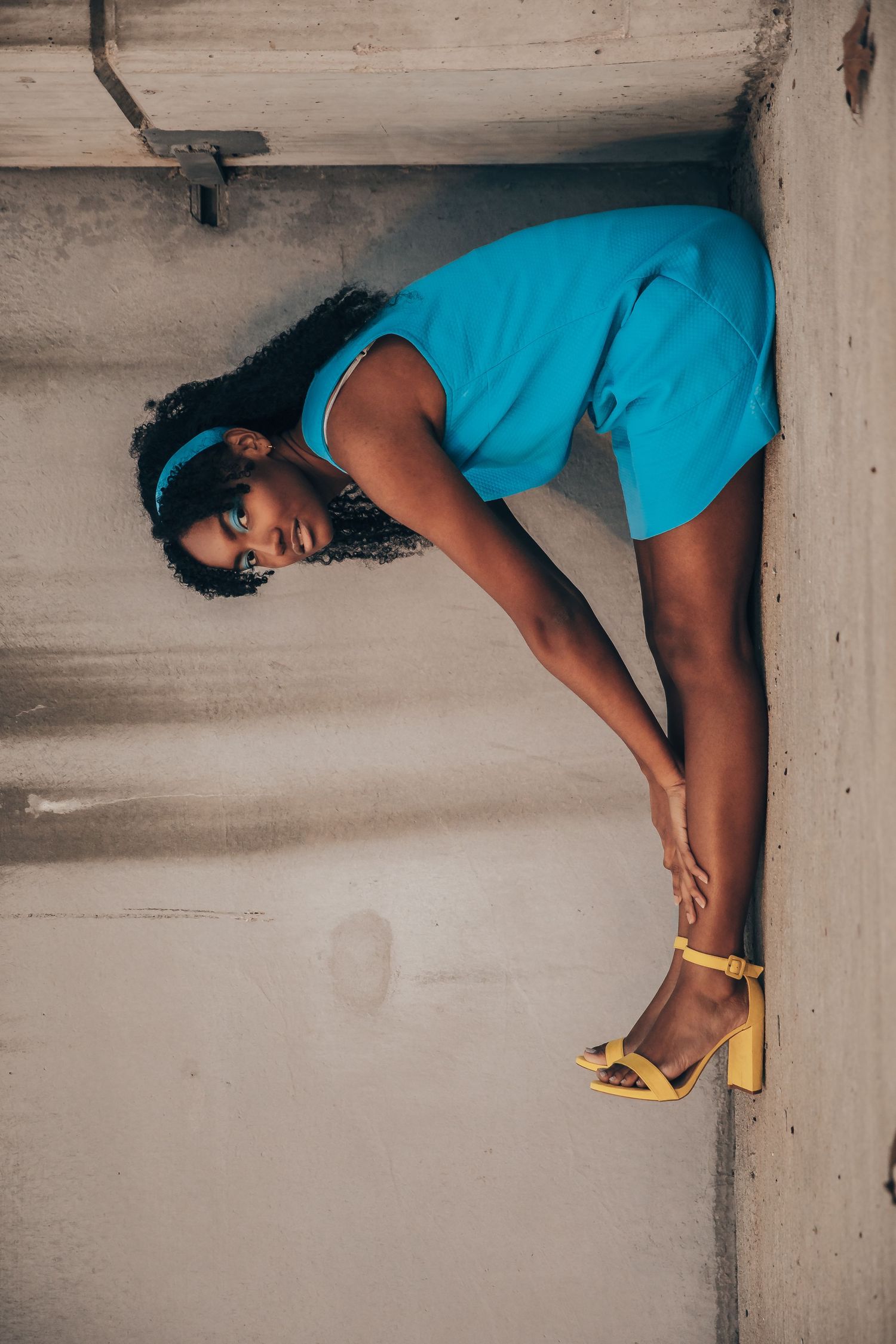Woman in blue romper and yellow heels poses gracefully between concrete walls, wearing blue sunglasses and headband.