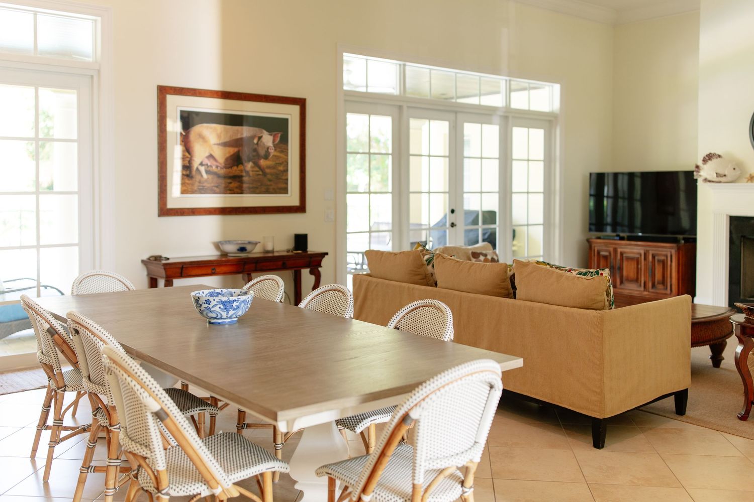 Bright open-plan dining and living room with white chairs, wooden table, beige sofas, and large French windows.