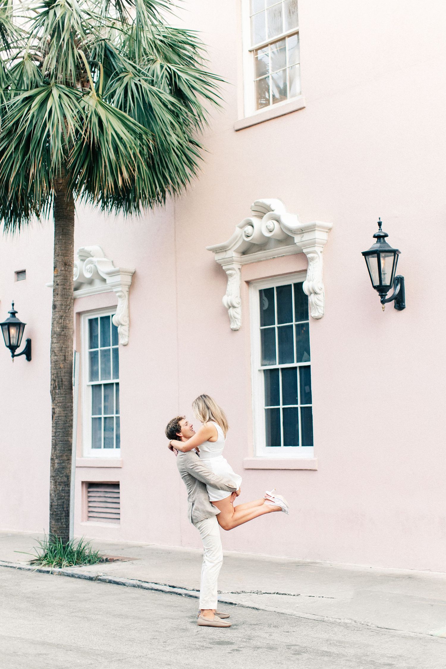 Romantic couple embracing as man lifts woman in front of a pink historic building with palm tree.