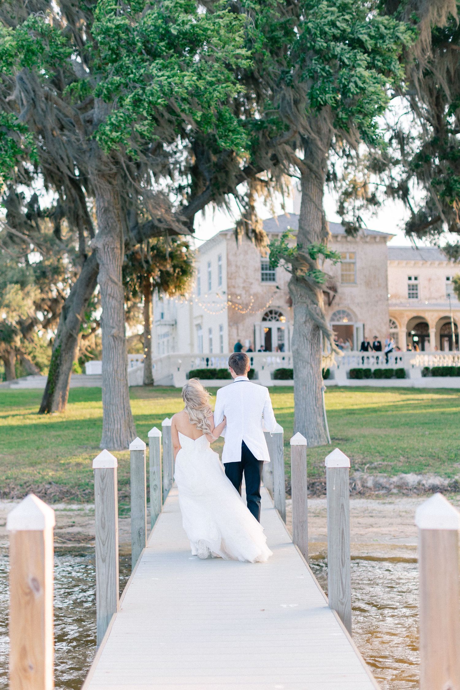Bride and groom walking hand in hand down a dock toward an elegant estate surrounded by lush trees, wedding at Bella Cosa