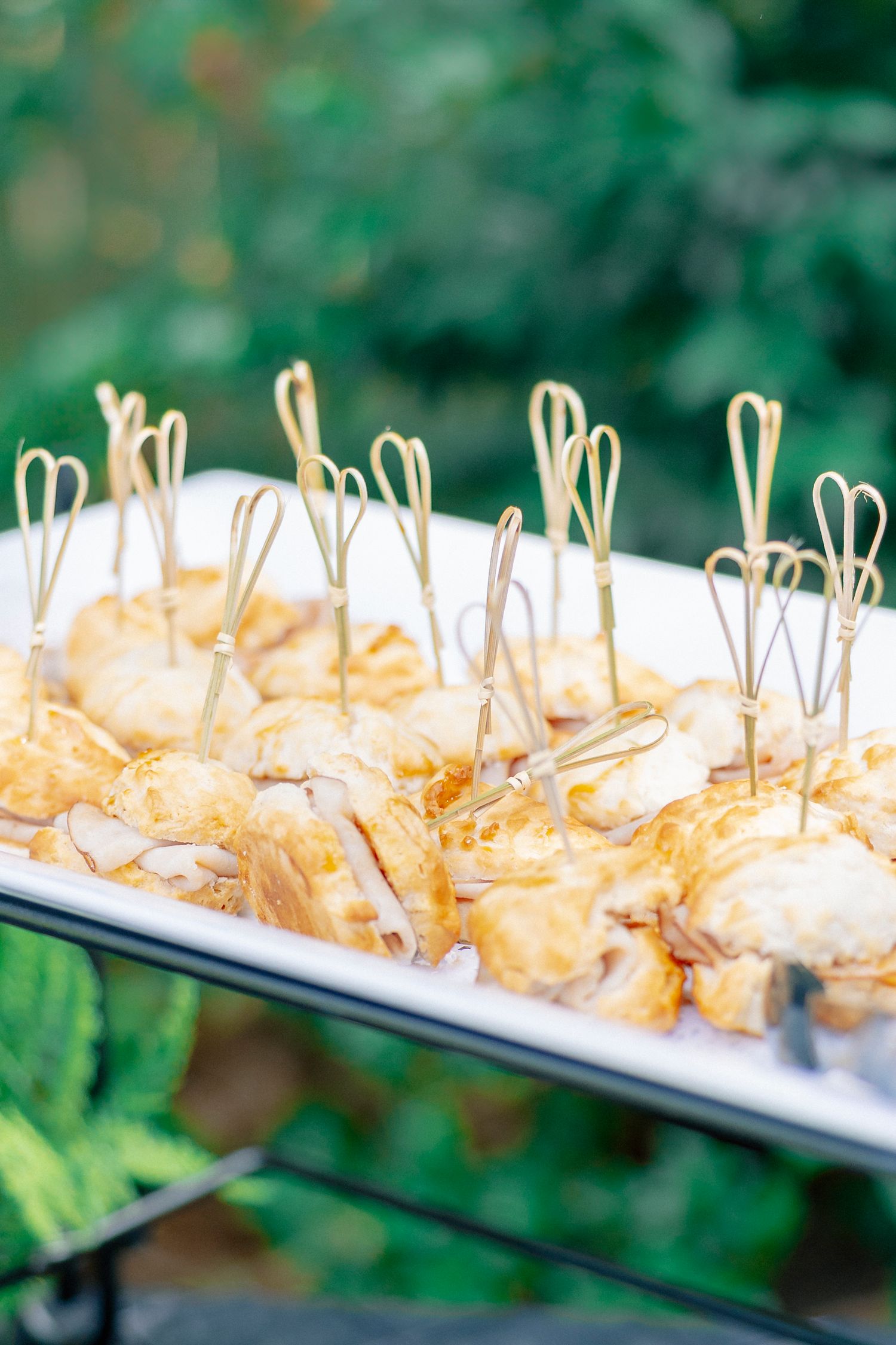 Golden appetizers on decorative picks arranged on a white tray at an outdoor catered event.