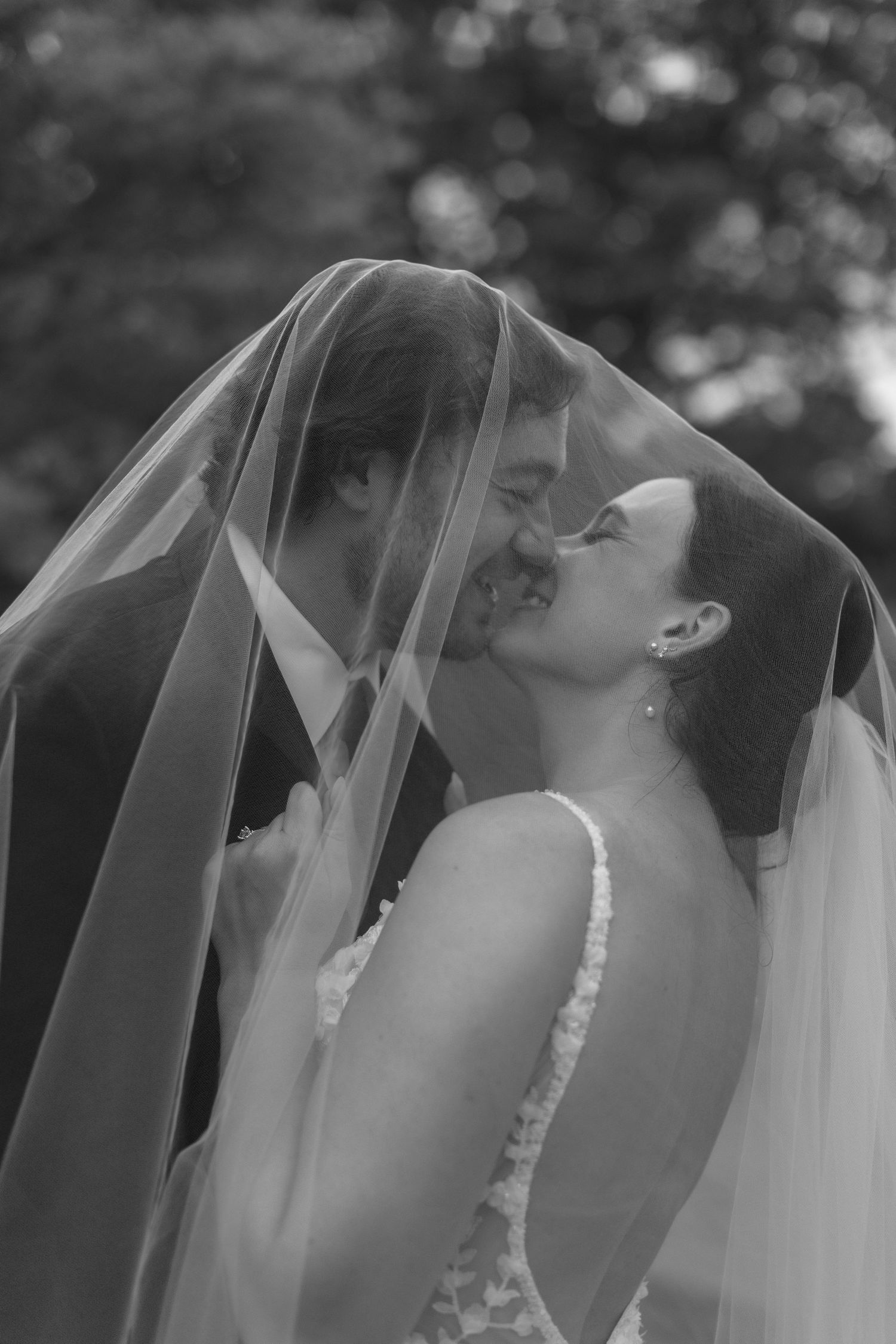 Black and white photo of bride and groom sharing a kiss under a flowing wedding veil outdoors.