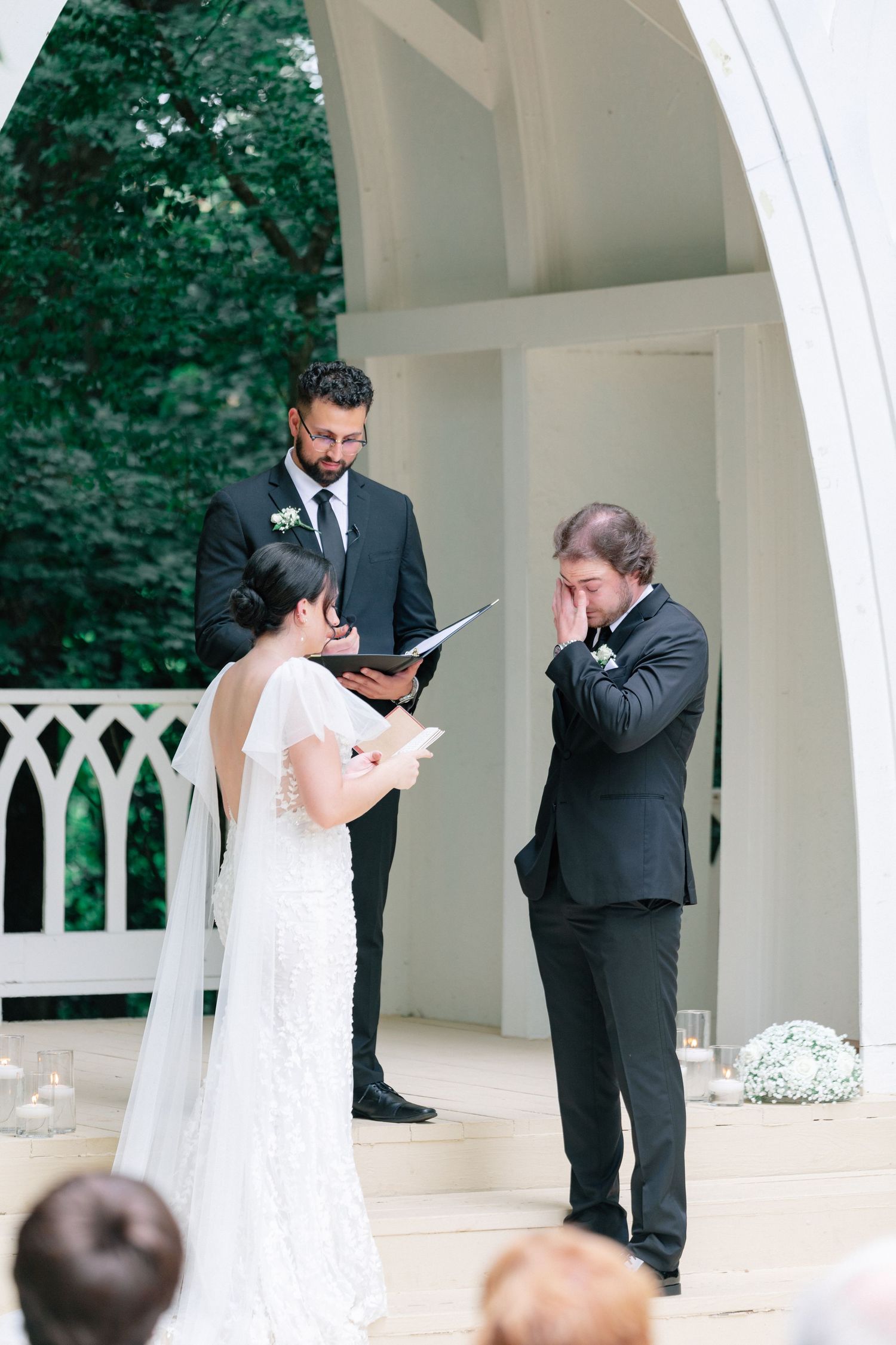 Groom wiping tears during outdoor wedding ceremony vows under white arch gazebo with officiant.