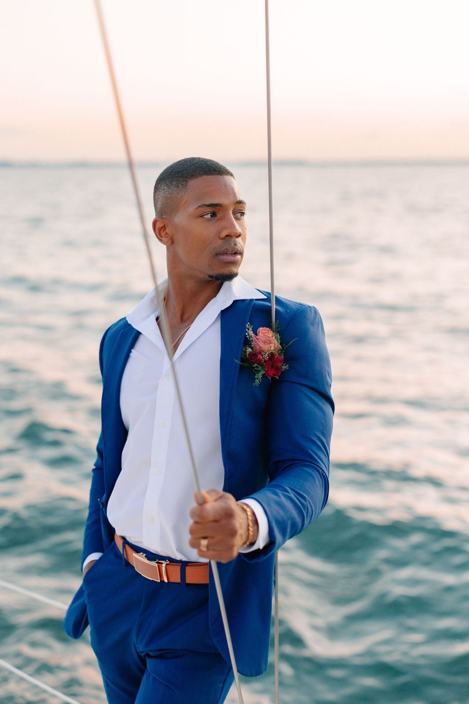 Stylish groom in blue suit with boutonniere standing on a sailboat at sunset over the ocean.
