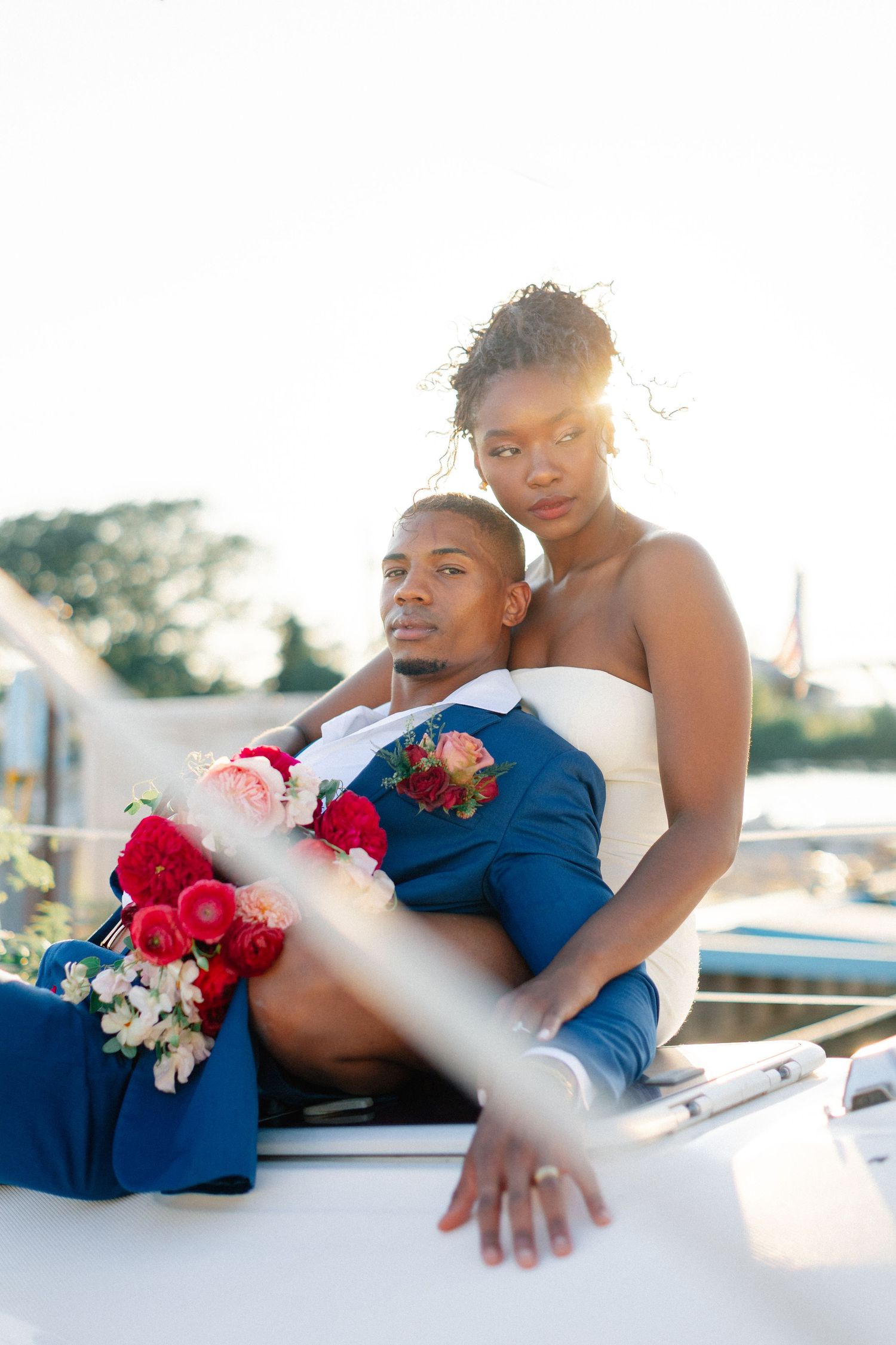 Bride and groom posing on a white yacht at sunset, holding a red and pink wedding bouquet.