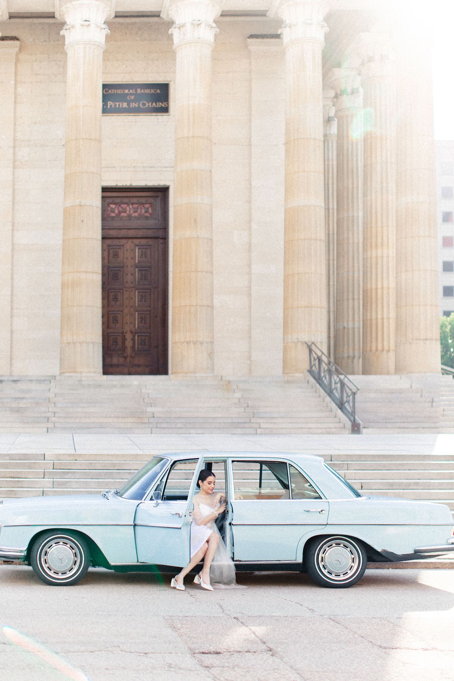 Bride in white dress leaning against vintage mint green Mercedes-Benz outside a neoclassical building with tall columns.
