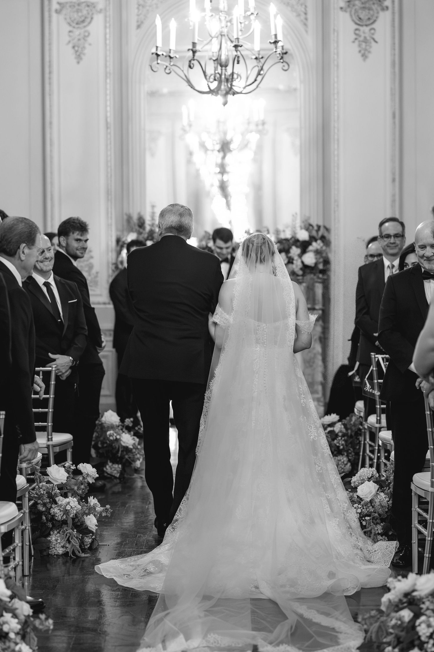Black and white photo of a bride walking down the aisle with her escort in an elegant chandelier-lit wedding venue.