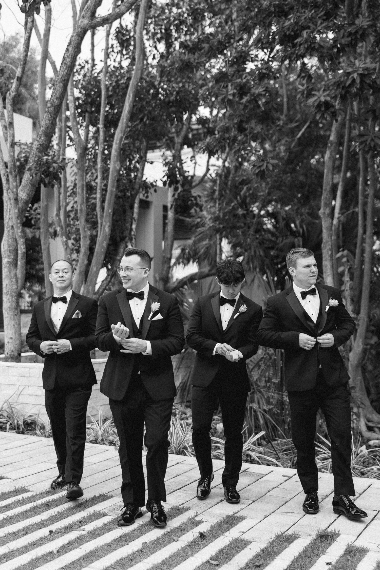 Four groomsmen in black tuxedos and bow ties walk together outdoors in a black and white wedding photo.