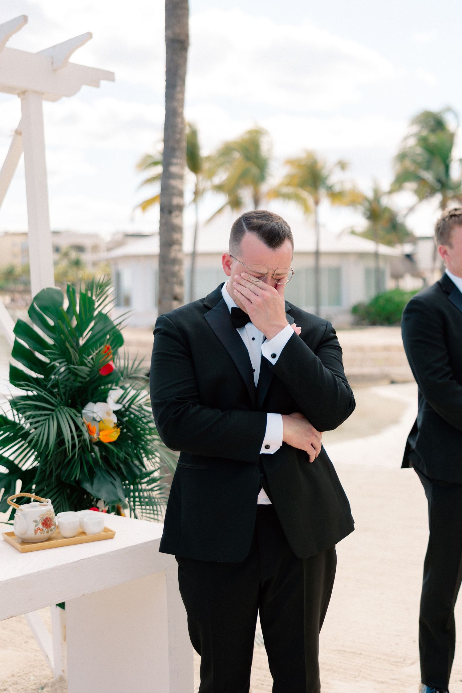 Emotional groom in black tuxedo covering his face at outdoor beach wedding ceremony with tropical floral decorations.