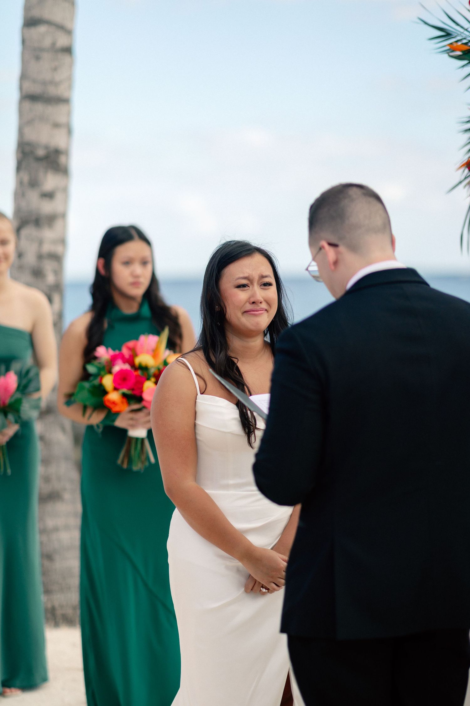 Bride crying happy tears during beach wedding ceremony with bridesmaids in green dresses holding colorful bouquets.