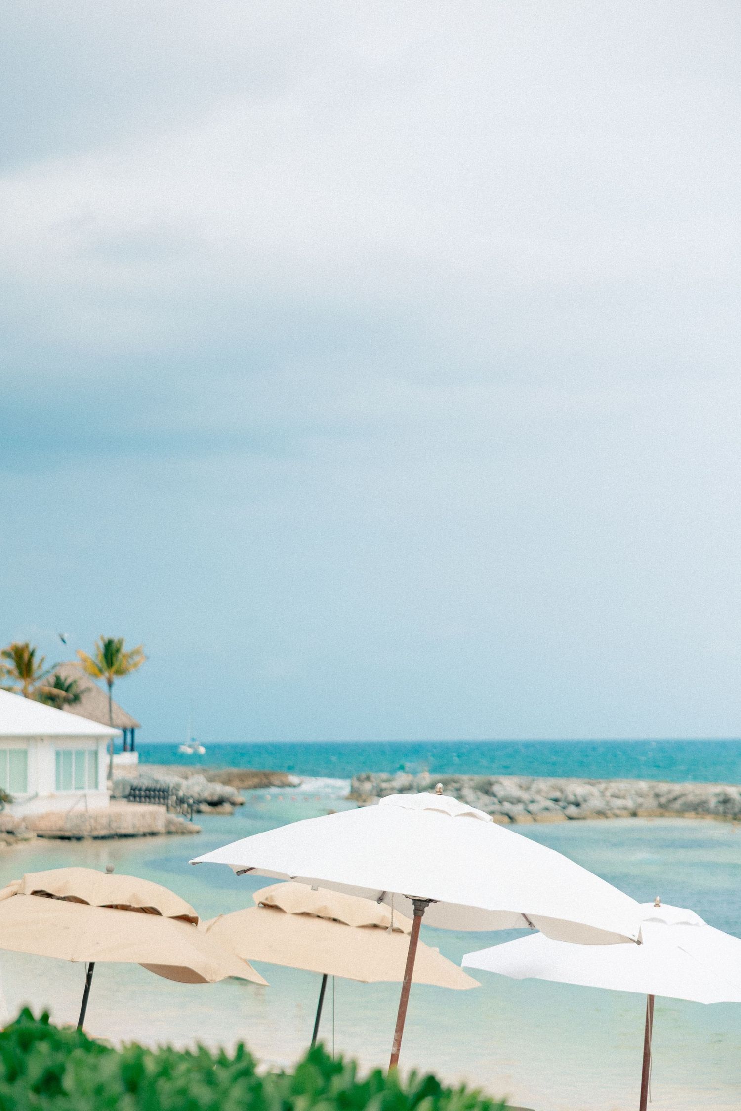 White beach umbrellas line a serene tropical resort shoreline with turquoise waters and palm trees under a cloudy sky.