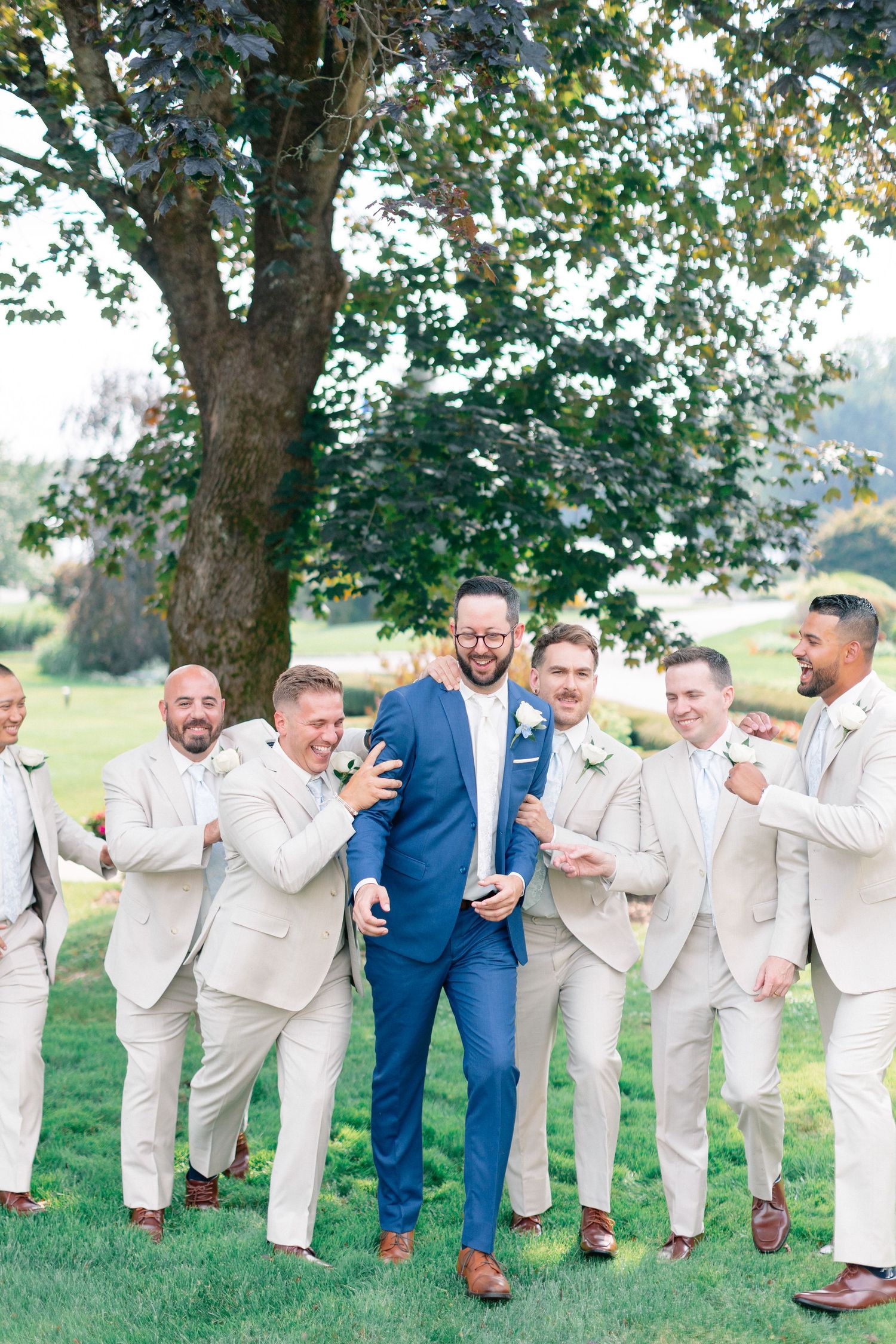 Groom in blue suit laughing with groomsmen in tan suits outdoors under a large tree on a sunny wedding day.
