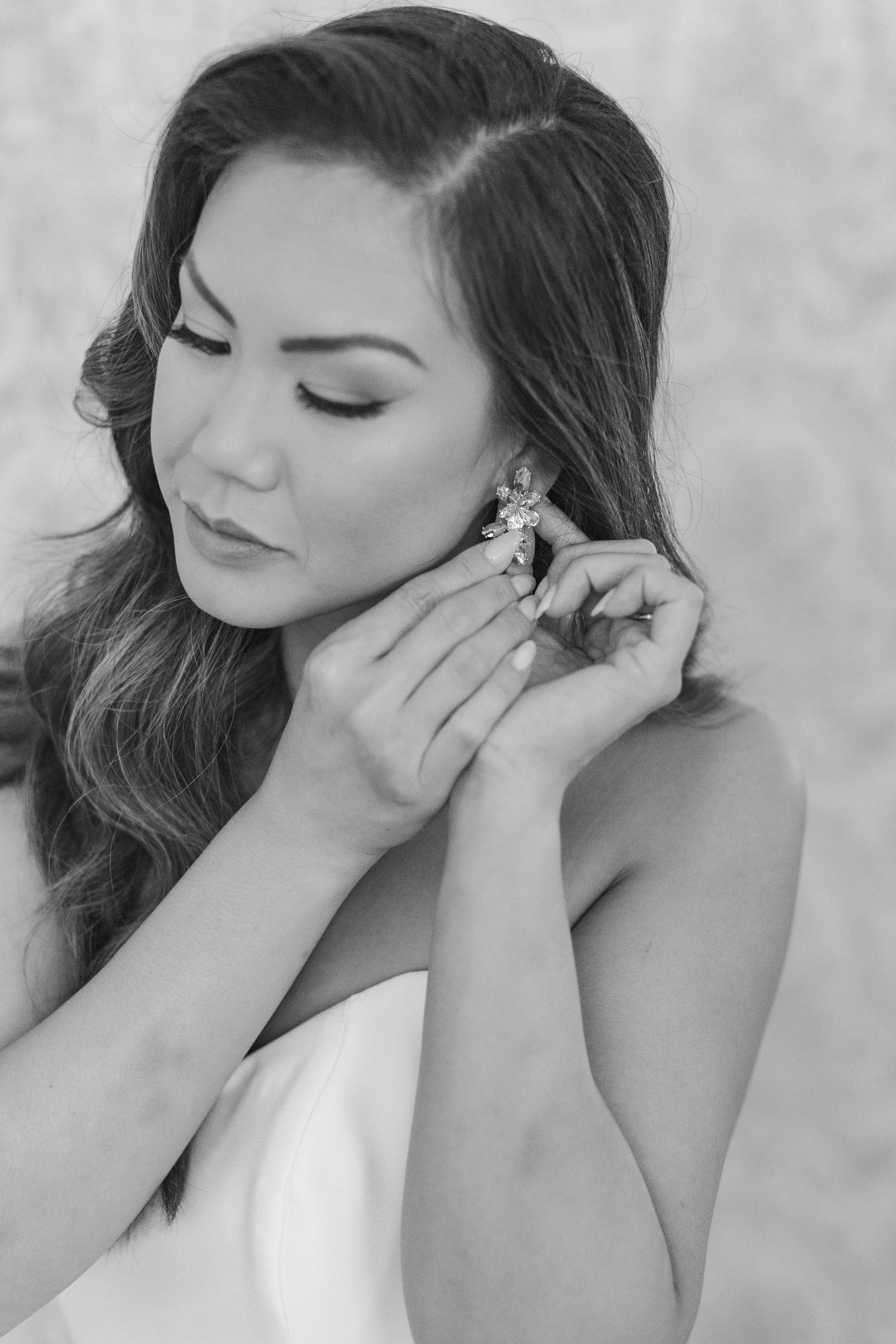 Bride in white dress putting on crystal earring, elegant bridal beauty portrait in black and white.