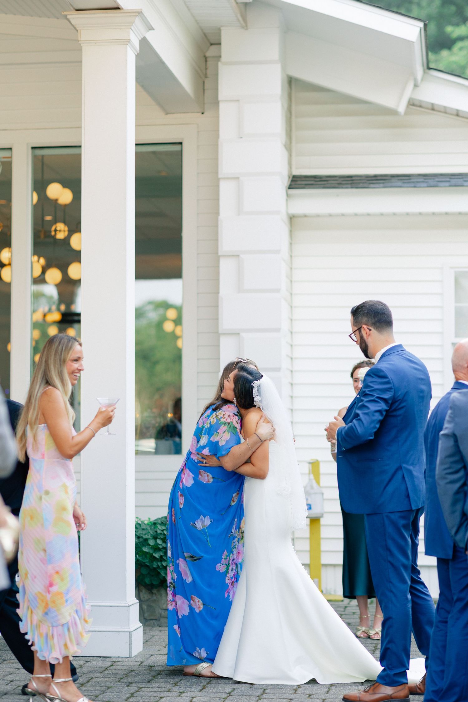 Bride in white gown hugging guest in floral blue dress outside elegant white venue during wedding reception.