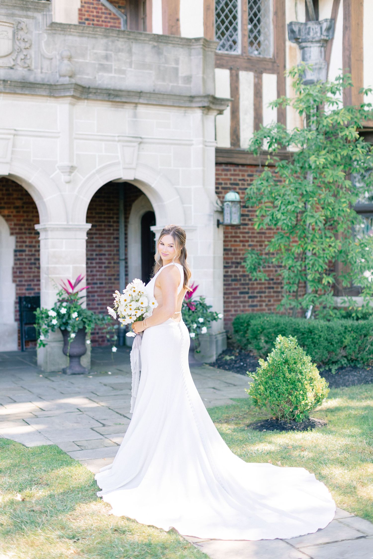Bride in elegant white gown with train holds bouquet, posing before a brick manor with arched stone entrance.