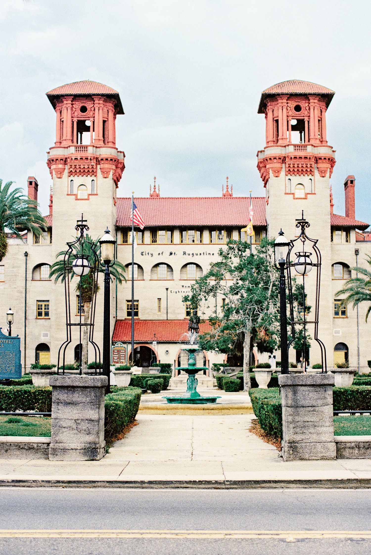 Historic City of St. Augustine building with red towers, manicured gardens, fountain, and American flag under cloudy skies.