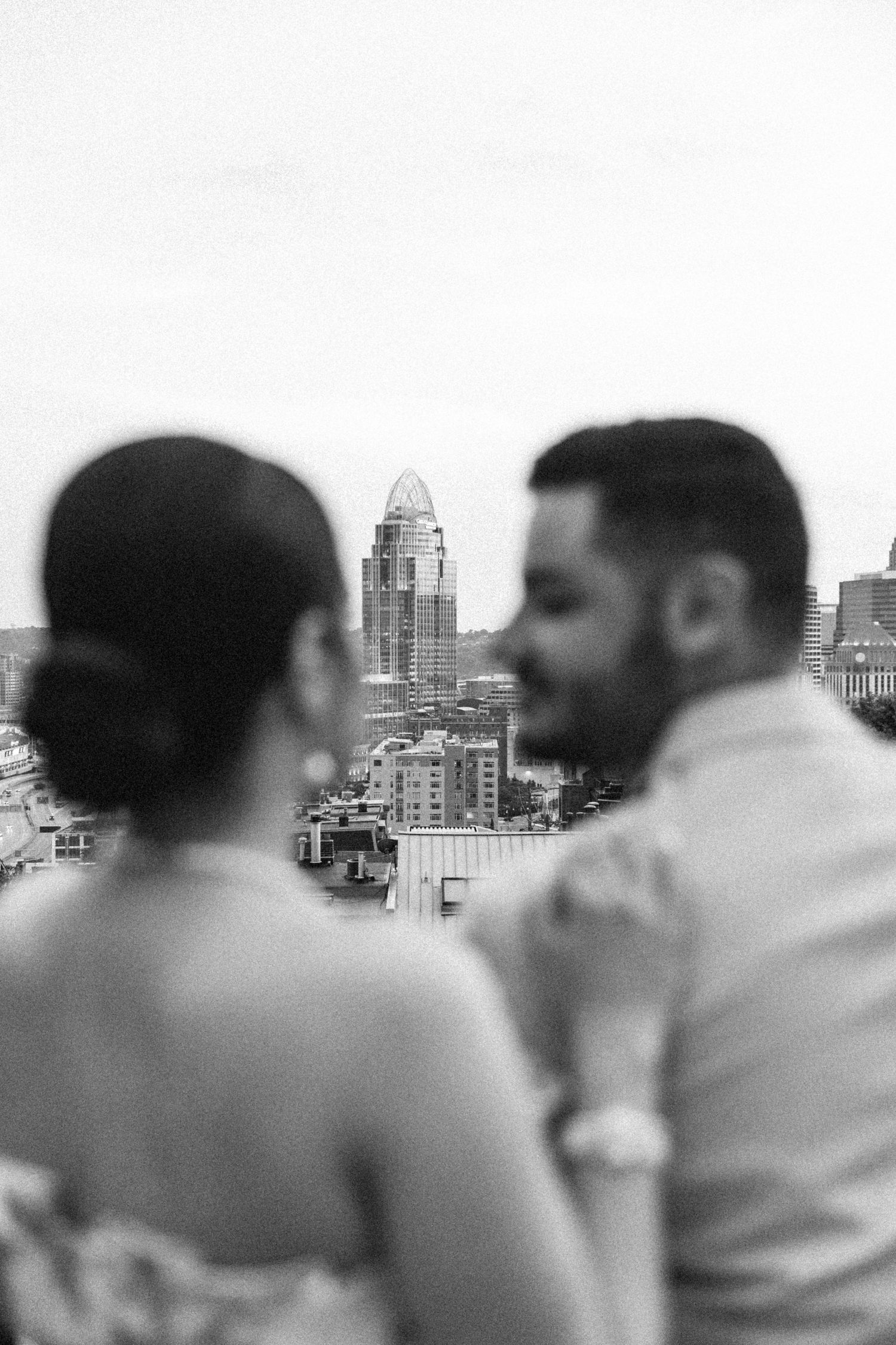 Black and white photo of couple with blurred city skyline and tall skyscraper in background.