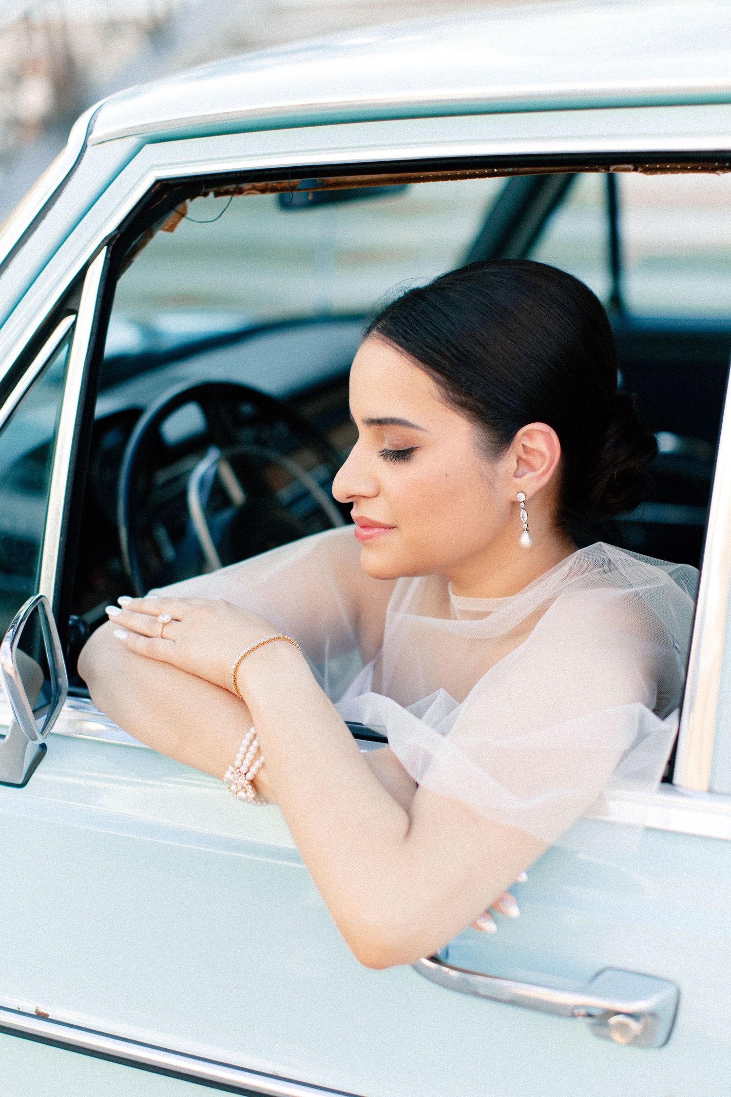 Elegant bride in sheer dress leaning out of vintage car window, wearing pearl bracelet and drop earrings.