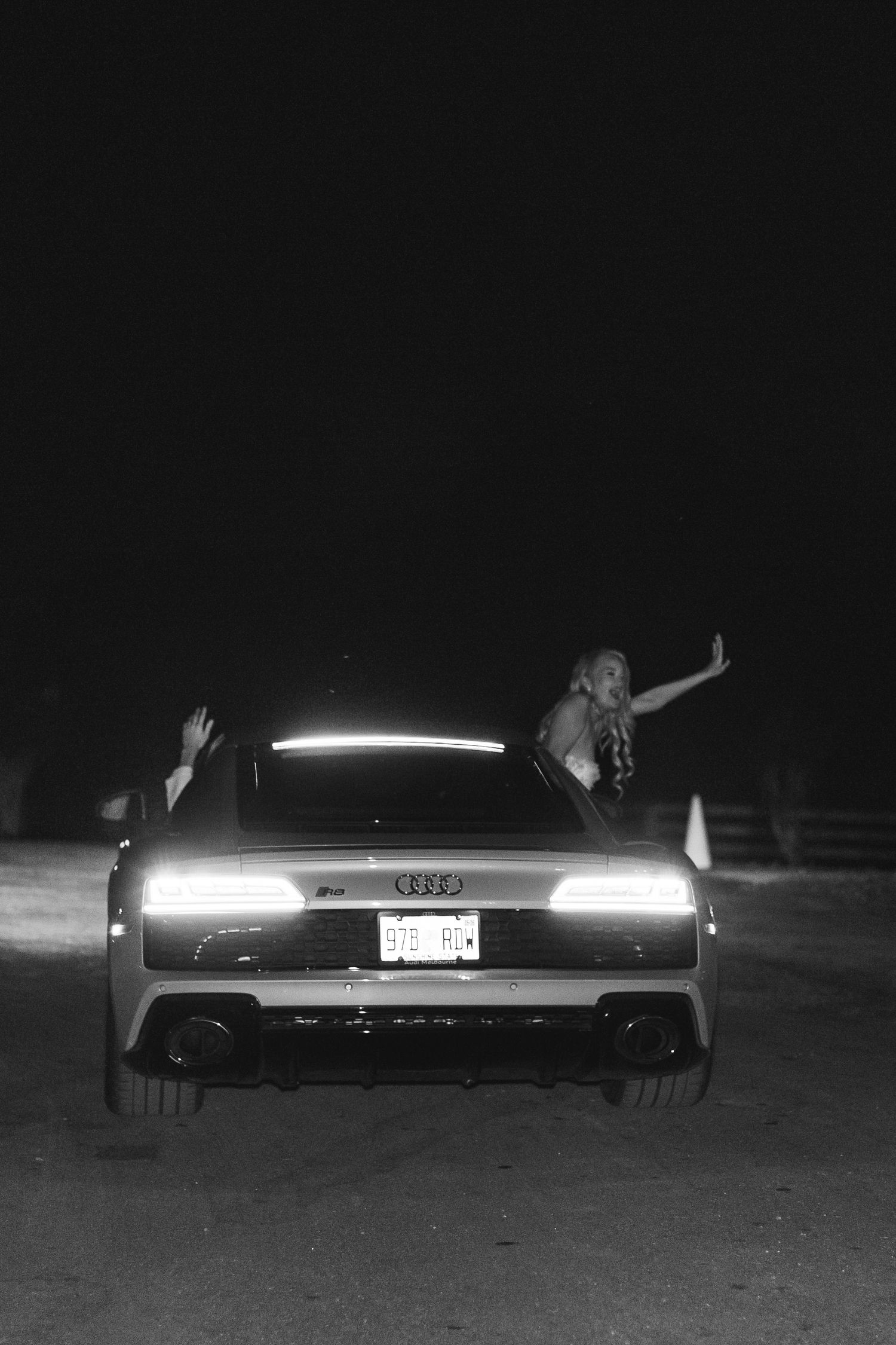 Black and white photo of two people posing behind an Audi car at night with headlights illuminated, wedding at Bella Cosa