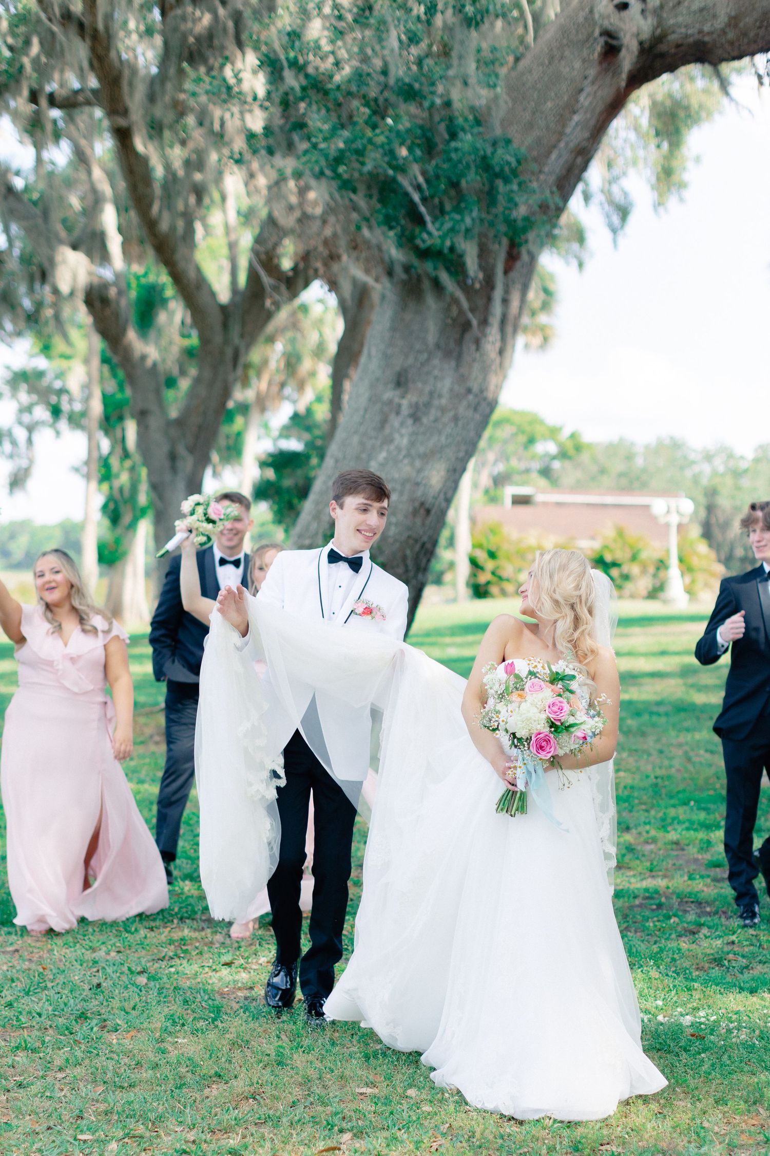 Wedding party walks outdoors under large oak trees, bride holding pink bouquet in white gown, groom in white tuxedo, wedding at Bella Cosa